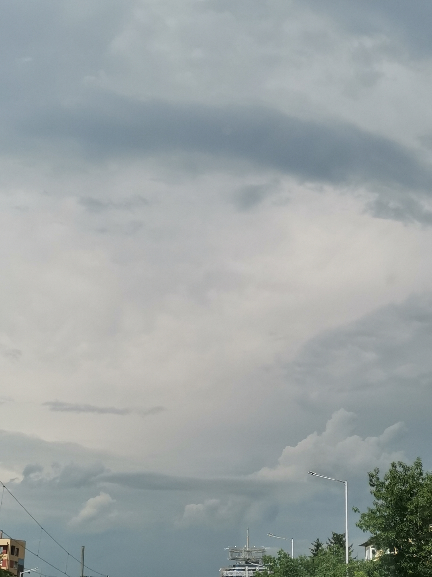The image shows a cloudy sky over an urban environment. In the foreground, there are several buildings and trees. A streetlamp is visible on the right side of the image. In the background, there are more buildings and a large communication tower. The sky is filled with dark, heavy clouds, suggesting an approaching storm. The lighting is dim and diffused. The scene appears to be a street view in a city, possibly in Sofia, Bulgaria, given the presence of specific architectural styles and vegetation.