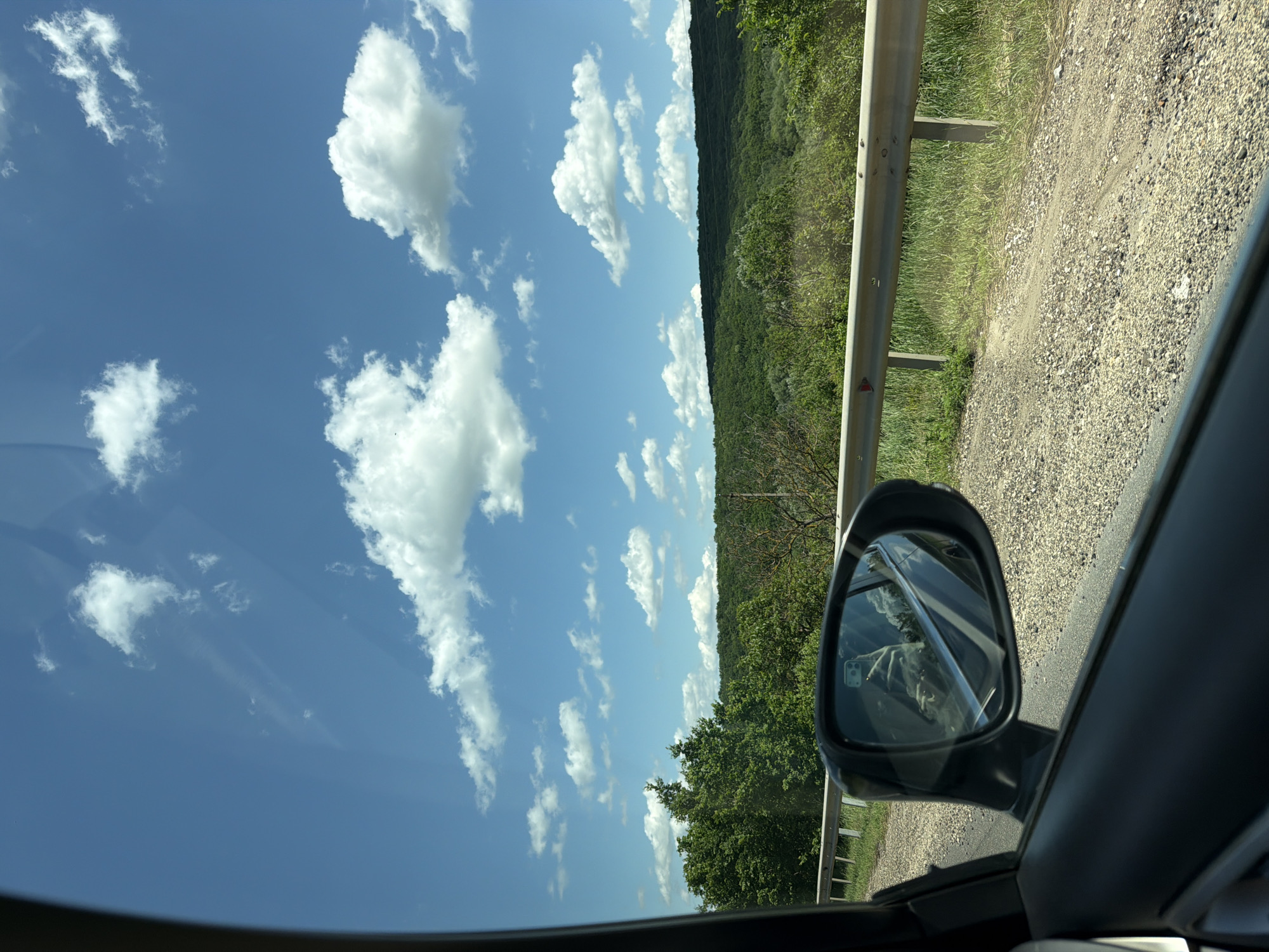 The image shows a view from inside a car in Peresecina, Moldova. The scene captures a sunny day with a blue sky dotted with fluffy white clouds. In the foreground, the car's side mirror reflects the interior of the car and the driver holding a phone. A guardrail runs along the side of a gravel road, bordering a lush green landscape of rolling hills covered in trees. The foliage is dense and vibrant, indicative of the summer season. The road appears unpaved or poorly maintained, with loose gravel and dirt. The overall impression is one of a rural drive on a clear, bright day.