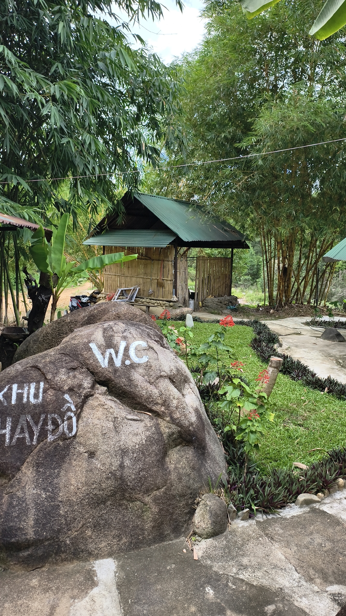 In Khanh Vinh, Vietnam, a natural setting features a bamboo hut with a green metal roof and bamboo walls, partially obscured by lush green bamboo trees. In the foreground, a large boulder is marked with white paint, displaying "W.C." on its upper section and "KHU HAYDO" on its lower section. The surrounding area is landscaped with green grass and small red flowers, with concrete pathways winding through the greenery. A motorbike is parked near the hut.