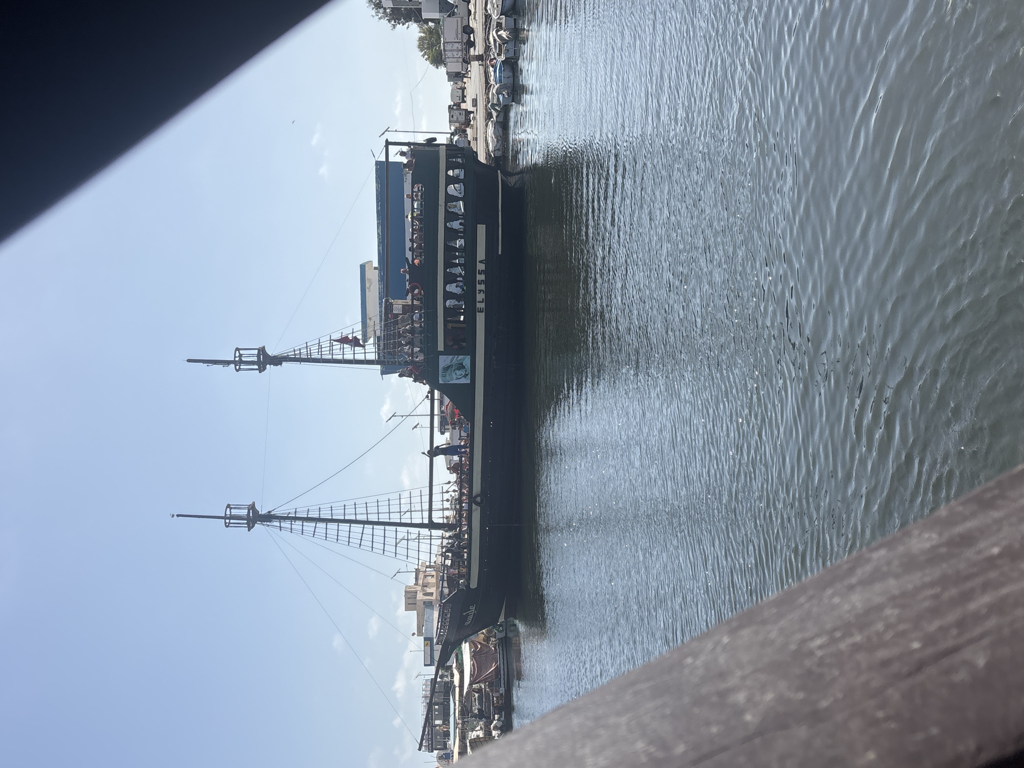 A dark green pirate-style tour boat named "ELYSSA" is docked in the harbor of Houmt Souk, Tunisia. The boat, which has two masts and appears to be carrying passengers on its upper deck, is positioned in the foreground. The water in the harbor is calm with small ripples. In the background, other boats are docked, and buildings, including a light blue structure, are visible along the waterfront. The sky is clear and blue with a few faint clouds. The scene is observed from a slightly elevated perspective, with a dark, out-of-focus wooden railing in the lower left corner of the frame.