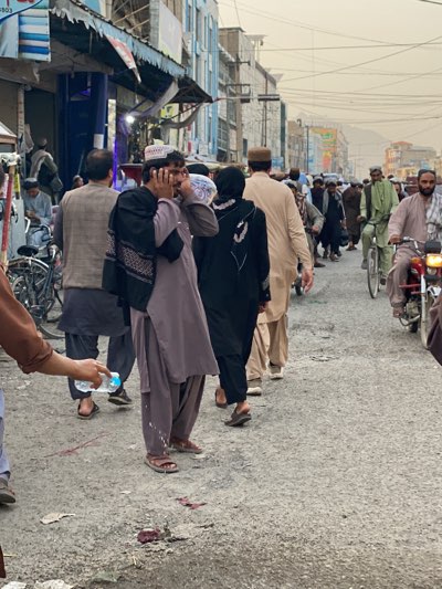 The image depicts a street in Kandahar, Afghanistan, crowded with people. Many men are wearing traditional clothing, including shalwar kameez and head coverings. One man is holding his hands to his ears. A woman in a black abaya is walking away. There is a bicycle and a motorcycle in the street. The road surface is rough and stained, apparently with blood. A person is holding a plastic bottle, potentially to clean or wash away the blood. The buildings lining the street have awnings and appear to be shops. The sky is hazy and overcast. Overhead are many wires, possibly electrical.