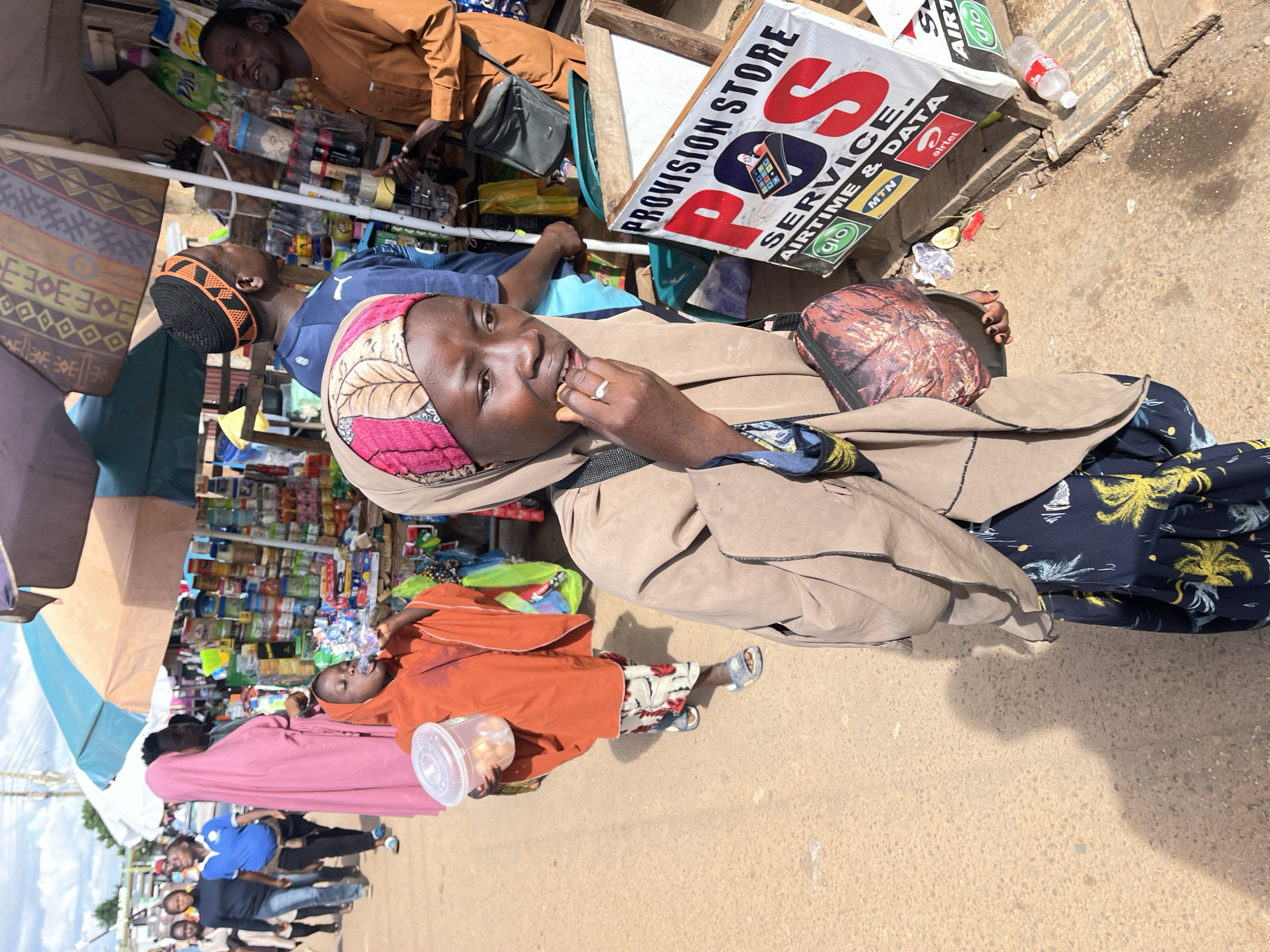 The image captures a street scene in Gura Top, Nigeria, with a provision store on the right side of the frame. A young girl, wearing an orange outfit and a headscarf, is in the foreground, holding a clear plastic container with small round items. She is looking to her right and has her hand to her mouth. Beside her, another person in a beige hijab is partially visible on the right. In the background, several individuals are walking along the dirt road, including a woman in a pink hijab, a man in a blue shirt and a cap, and another man in a brown outfit. The provision store displays various products and a sign that reads "PROVISION STORE POS SERVICE AIRTIME & DATA". The ground is sandy and there are scattered items, including a plastic bottle and debris near the store. The sky above is partly cloudy.