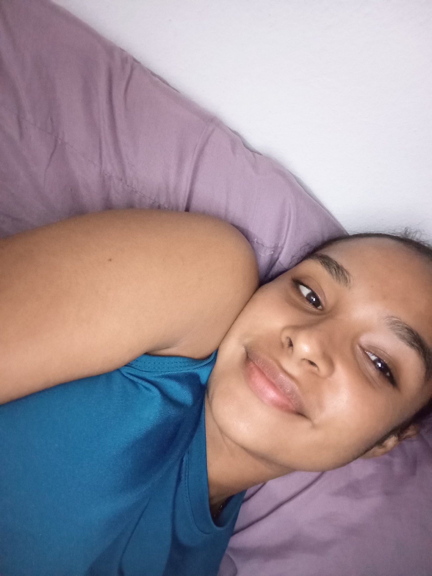 A young woman is captured in a close-up portrait, lying down and looking directly at the camera with a gentle, relaxed smile. She has medium-brown skin, dark eyes, and her dark hair is pulled back. She is wearing a teal or blue t-shirt or tank top, which is visible across her chest and shoulder. Her left arm, with a small mole on the bicep, is partially visible, appearing to be tucked under her head or resting on the pillow.
The setting appears to be indoors, likely a bedroom or living space, as she is resting on a soft lavender or light purple pillow or bedding. A plain white wall is visible in the upper right background, suggesting a simple interior. The lighting is soft and even, illuminating her face without harsh shadows, which could indicate indoor ambient light during the day or evening. The overall mood conveyed by her expression is calm and content. No specific outdoor elements or unique indoor decorations are visible to firmly establish the location, other than the provided context of Juan Griego, Venezuela. There is no visible text in the image.