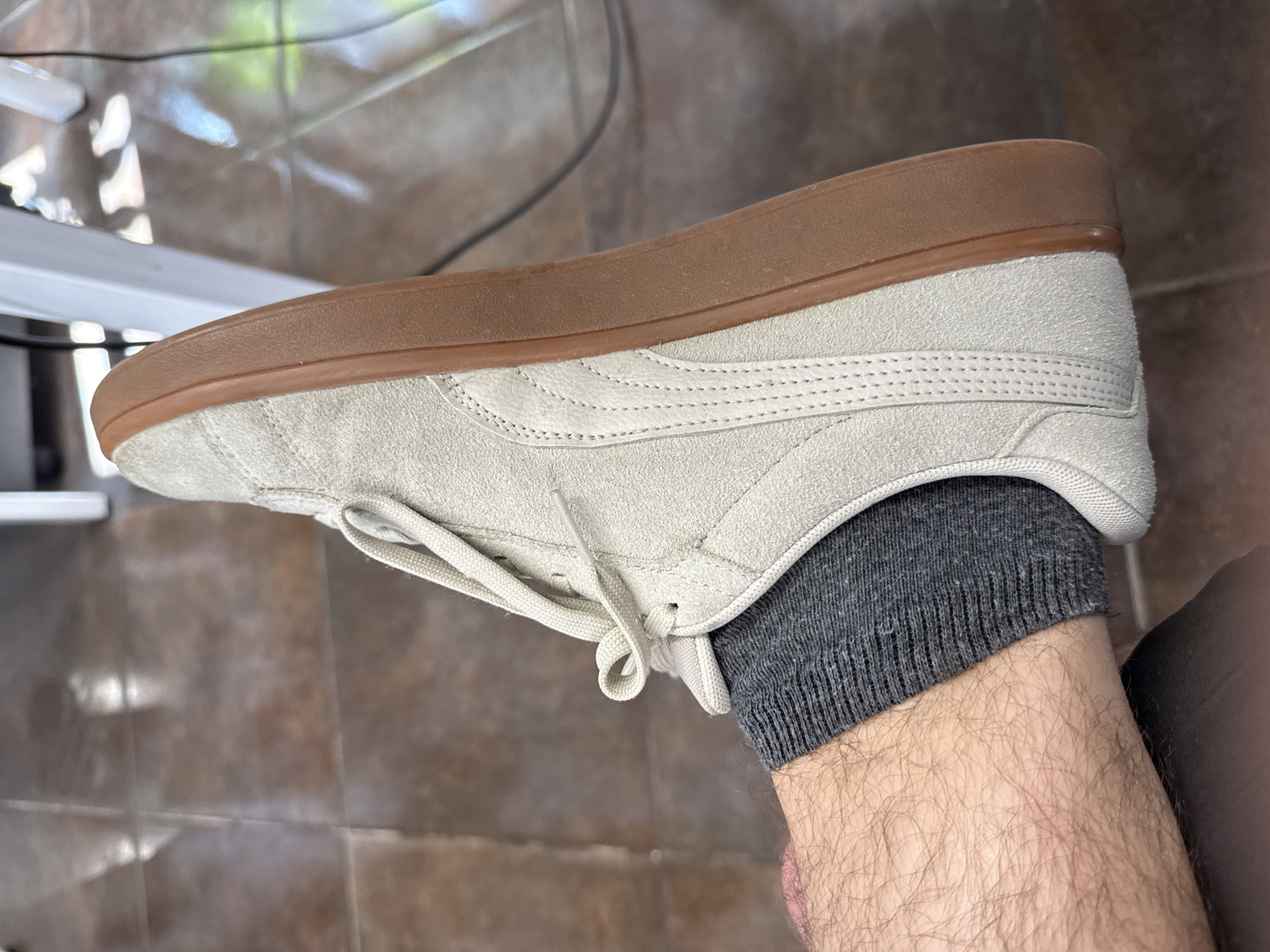 The image shows a close-up view of a person's right foot wearing a light-colored, low-top sneaker with a gum sole and dark gray socks. The sneaker is made of suede and has white laces. The person's lower leg, covered in dark hair, is visible below the sock. The background consists of a tiled floor and a blurred reflection of what appears to be a desk or table, with some greenery visible in the reflection. The lighting suggests it is daytime. The context provided indicates the location is Łódź, Poland. No text is visible in the image. The scene captures a mundane moment, with no specific activity or event clearly depicted beyond the act of wearing the footwear.
