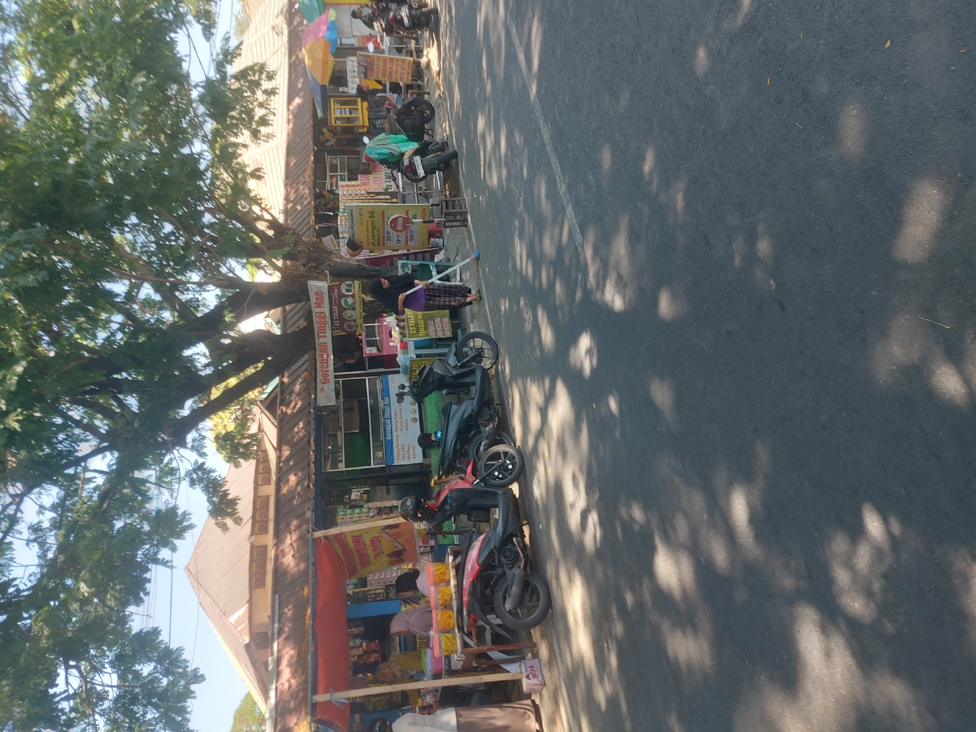 The image depicts a street scene in Makassar, Indonesia, likely a roadside market or a row of small shops. The foreground shows a paved road with tree shadows dappled across it. In the mid-ground, a row of small food stalls or shops lines the street, identifiable by their awnings and displays of goods. Several motorcycles are parked along the roadside. A woman in a black headscarf and patterned pants is sweeping the pavement near one of the stalls. Other individuals are visible behind the counters of the shops, appearing to be vendors. The architecture suggests a tropical climate, with open-air structures and large trees providing shade. The overall atmosphere is that of a busy local market or commercial area.