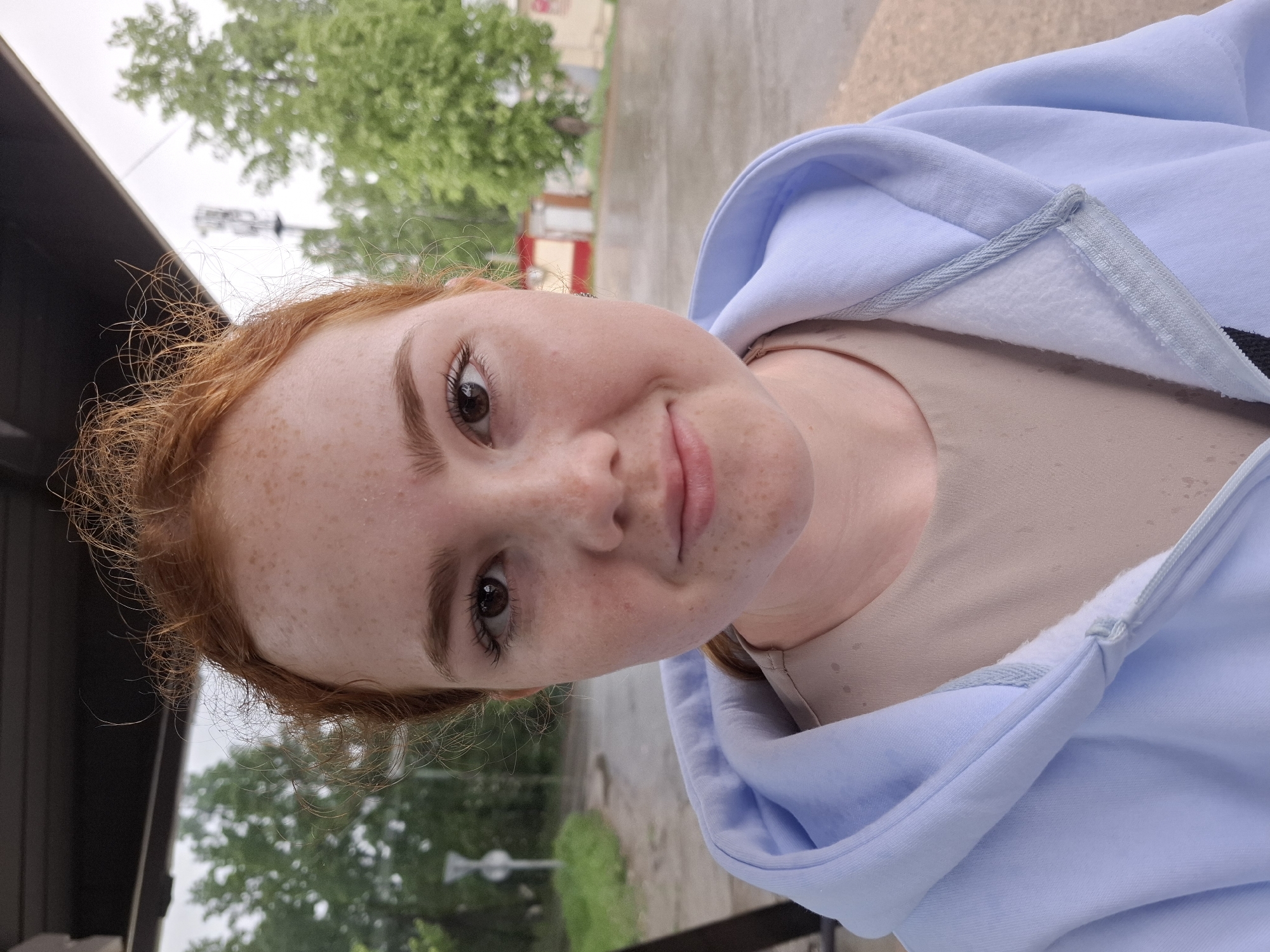 A close-up portrait shows a fair-skinned individual with red hair and freckles. The person is wearing a light blue hooded sweatshirt over a tan-colored shirt. The background reveals a blurry urban environment, suggesting an outdoor setting. The location is identified as Chaikovsky, Russia. The person is looking directly at the camera with a slight smile. The scene gives the impression of a casual, candid snapshot within an urban landscape.
