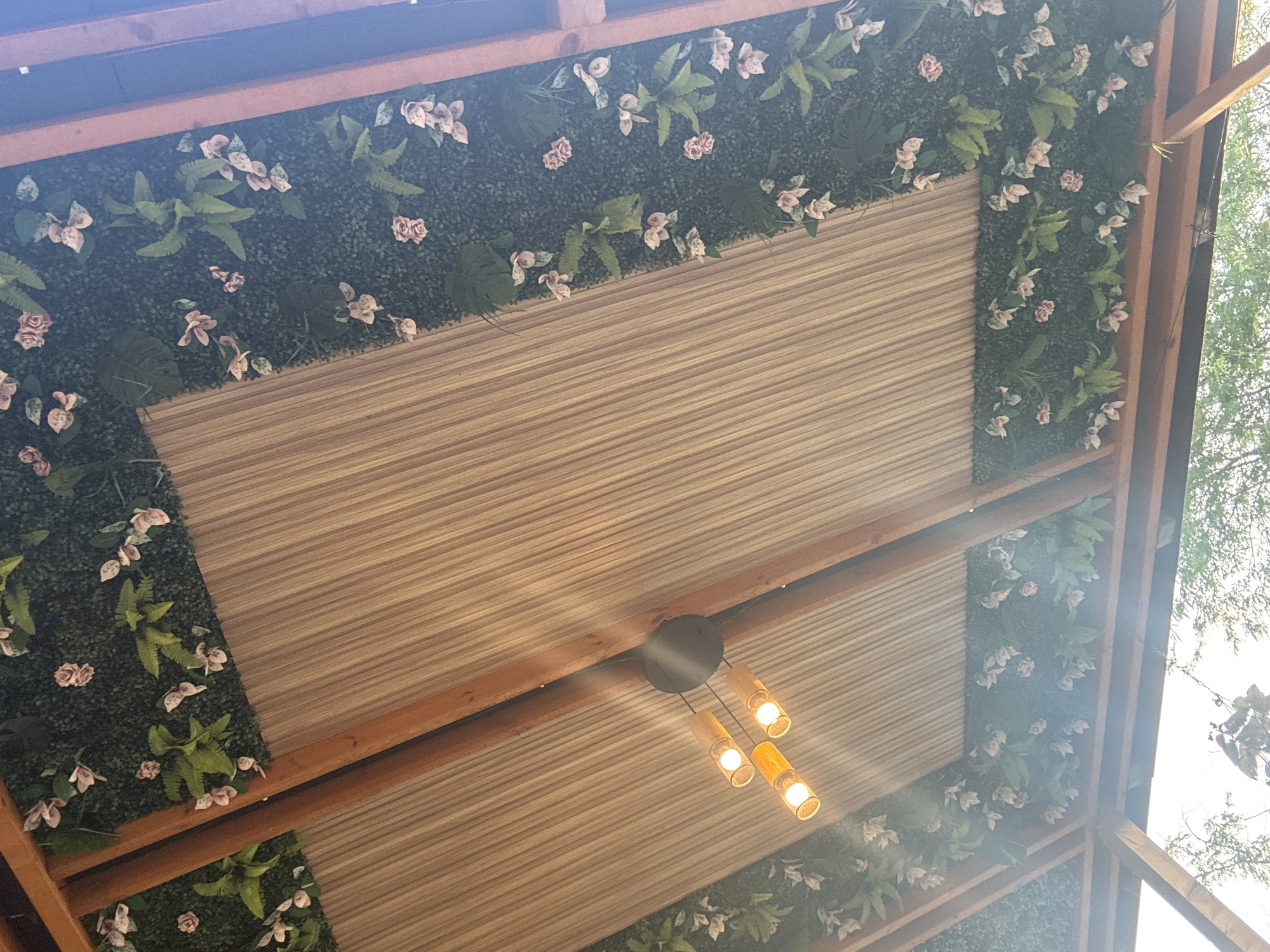 The image shows the ceiling of an outdoor structure, possibly a gazebo or pavilion, in Primorsko, Bulgaria. The ceiling is made of two sections of light brown, ribbed material, supported by wooden beams. The edges of the ceiling are adorned with artificial greenery, interspersed with small pink and white flowers and some fern-like foliage. A modern light fixture with three cylindrical lamps hangs from the center of one of the ceiling sections, emitting a warm, yellow light. The structure appears to be open to the sky, with hints of trees and foliage visible in the lower part of the frame, suggesting an outdoor setting. The overall impression is of a decorative and possibly sheltered outdoor seating or gathering area.