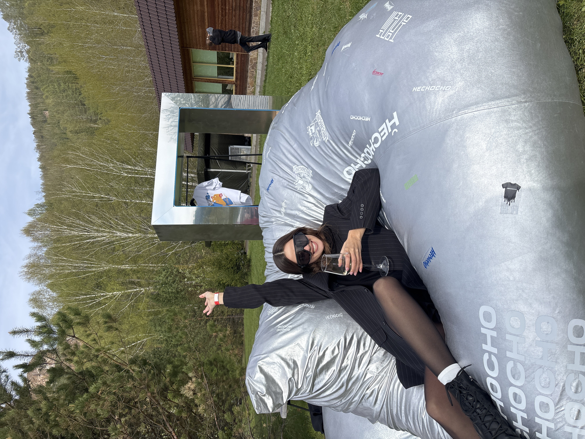 Here is a detailed description of the image:
**Scene Overview:** The image shows a woman reclining on a large, inflated silver surface with the word "HECHOCHO" repeatedly printed on it. The location appears to be outdoors, with trees and a building visible in the background.
**People:**
- A woman is reclining on the silver surface. She is wearing a dark pinstripe suit, dark sunglasses, and tights. She is holding a wine glass in one hand and has her other arm outstretched.
- A second person is visible in the background near a building, but they are distant and details are indistinct.
**Objects:**
- A large, inflated silver surface with "HECHOCHO" printed on it.
- A wine glass.
- A dark pinstripe suit.
- Dark sunglasses.
- Tights.
- A rectangular metal frame is behind the woman.
- A white shirt is displayed in the metal frame.
- Trees and foliage are in the background.
- A wooden building is partially visible.
**Actions and Interactions:**
- The woman is reclining and holding a wine glass, appearing relaxed and posing for a photo.
- She is gesturing with one hand, as if directing someone or something.
**Environment:**
- The scene is outdoors, likely in a park or open space.
- Trees are in the background, suggesting a natural environment.
- There is a building in the background.
**Location:** It cannot be confirmed that the location is in the city of Krasnoyarsk, Russia. More information or metadata would be needed to confirm the exact location.