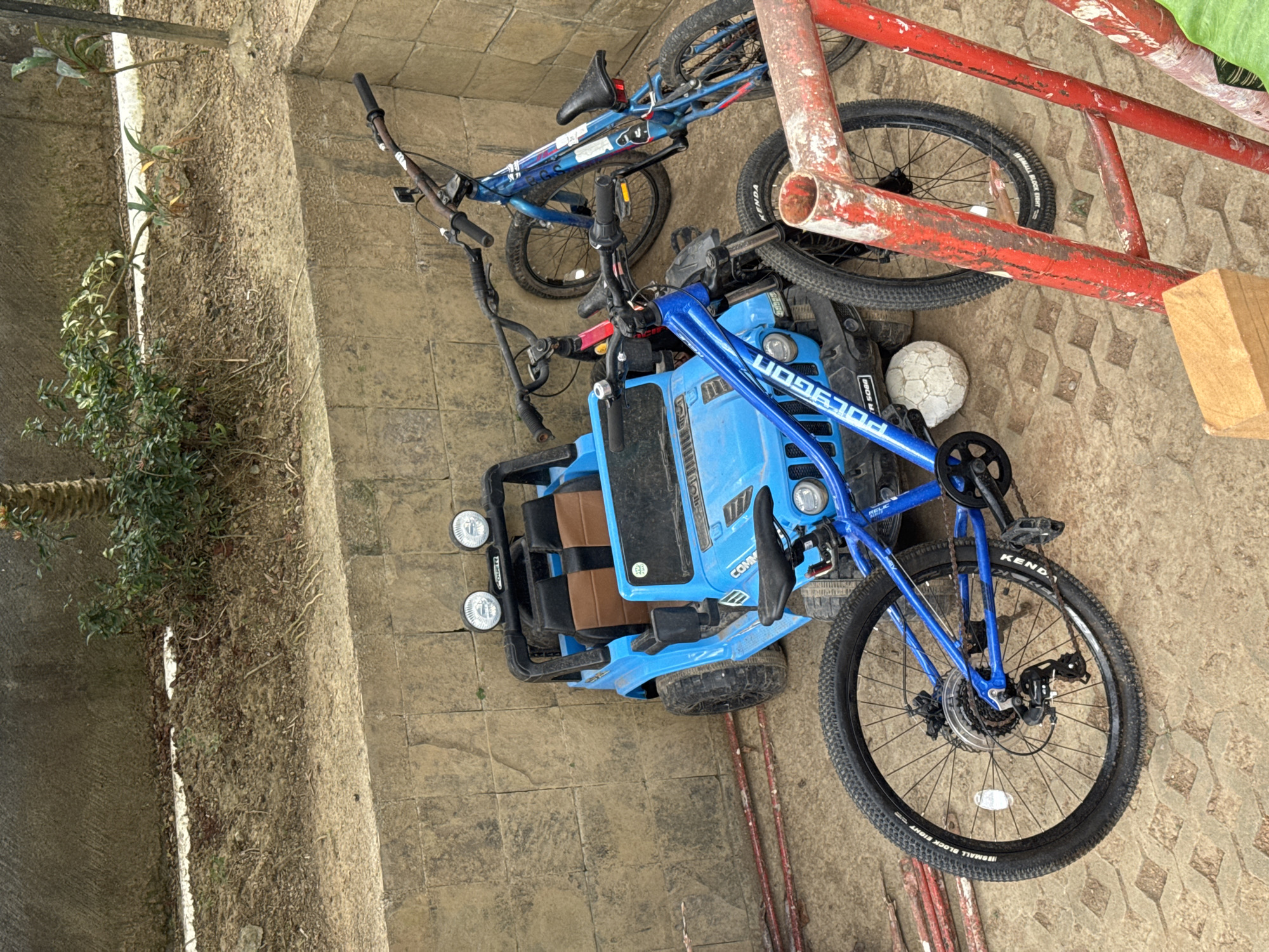 In Kuta Selatan, Indonesia, an outdoor scene features a blue toy jeep parked against a textured stone wall. Two blue bicycles are positioned in front of and beside the toy jeep. The foreground displays the rear wheel of one bicycle and a portion of a red metal structure. The ground is a paved surface with a geometric pattern. The environment appears somewhat unkempt, with dirt and vegetation visible behind the wall. No people are present in the image.