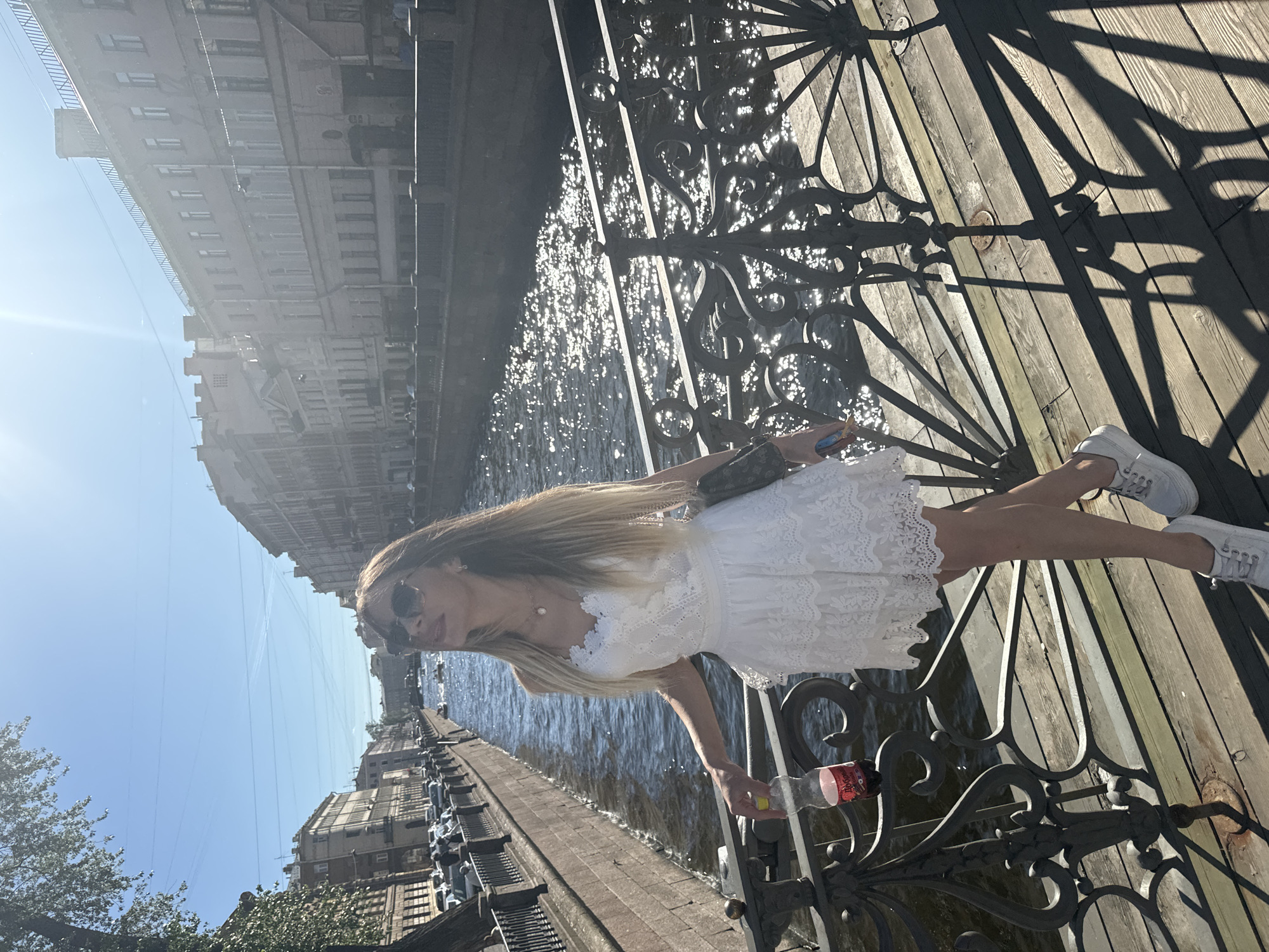 A young woman with long blonde hair stands on a wooden bridge, leaning against an ornate black metal railing. She is wearing a white lace dress and white sneakers. Her sunglasses are on, and she has a pearl necklace. She is holding a bottle of soda in her right hand. The bridge overlooks a canal, with buildings lining the opposite bank. The sun is shining, casting long shadows on the wooden planks of the bridge. The water in the canal reflects the sunlight, creating a shimmering effect. The sky is clear and blue. The scene appears to be set in Saint Petersburg, Russia, given the architectural style of the buildings and the presence of a canal. The overall mood is bright and summery, suggesting a pleasant day for a stroll. There is no visible text in the image.