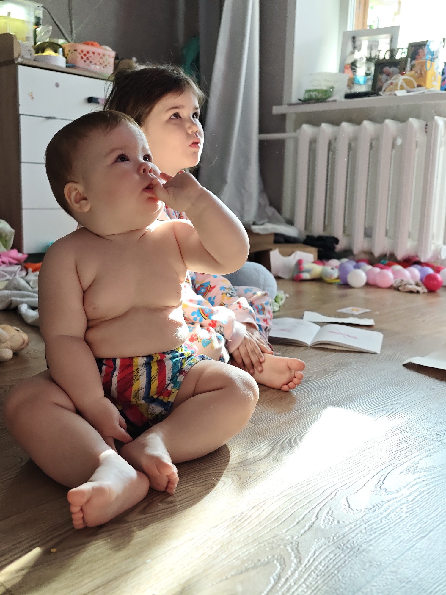 This image depicts two young children sitting on a wooden floor in a sunlit room, likely in Chaikovsky, Russia. In the foreground, a baby, possibly a toddler, sits with a chubby physique, wearing colorful striped shorts or a diaper cover. The baby looks upwards and slightly to the left, with fingers near its mouth, indicating focus or teething. Behind and to the right of the baby sits an older child with dark hair, also looking intently upwards and to the right, sharing a similar expression of concentration or curiosity.
The setting appears to be a child-friendly indoor space, possibly a living room or bedroom. Bright sunlight streams in from a window on the right side of the frame, creating illuminated patches on the light-colored wooden floor. The room shows signs of daily activity with various objects scattered around. To the left, part of a white dresser or cabinet is visible, holding a pink basket and other items. On the right, beneath the window, is a white radiator, a common feature in Russian homes, and on the windowsill above it are several framed photographs and other small items. A light-colored curtain hangs by the window.
On the floor, an open book lies near the older child's feet, and further back, a collection of colorful ball pit balls (white, pink, purple) is visible near the radiator. A stuffed animal can also be seen near the baby on the left. The children appear calm and engrossed in something outside the viewer's sight, suggesting they are watching a show, observing something outdoors, or engaging with another person in the room. The direct sunlight indicates it is daytime, likely morning or afternoon. There is no discernible text visible in the image.