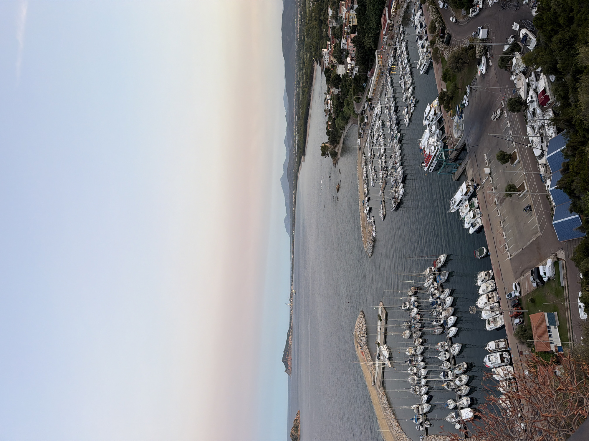 The image presents a high-angle view of a harbor filled with numerous sailboats and motorboats docked in calm water. The harbor is bordered by a town with buildings clustered along the coastline, surrounded by greenery. In the distance, a peninsula with a hill extends into the sea, and a range of mountains forms the backdrop. The sky transitions from a pale blue at the top to softer pinks and oranges near the horizon, suggesting either sunrise or sunset. The scene is static, with no visible human activity. The overall impression is one of a peaceful coastal town and its bustling marina.