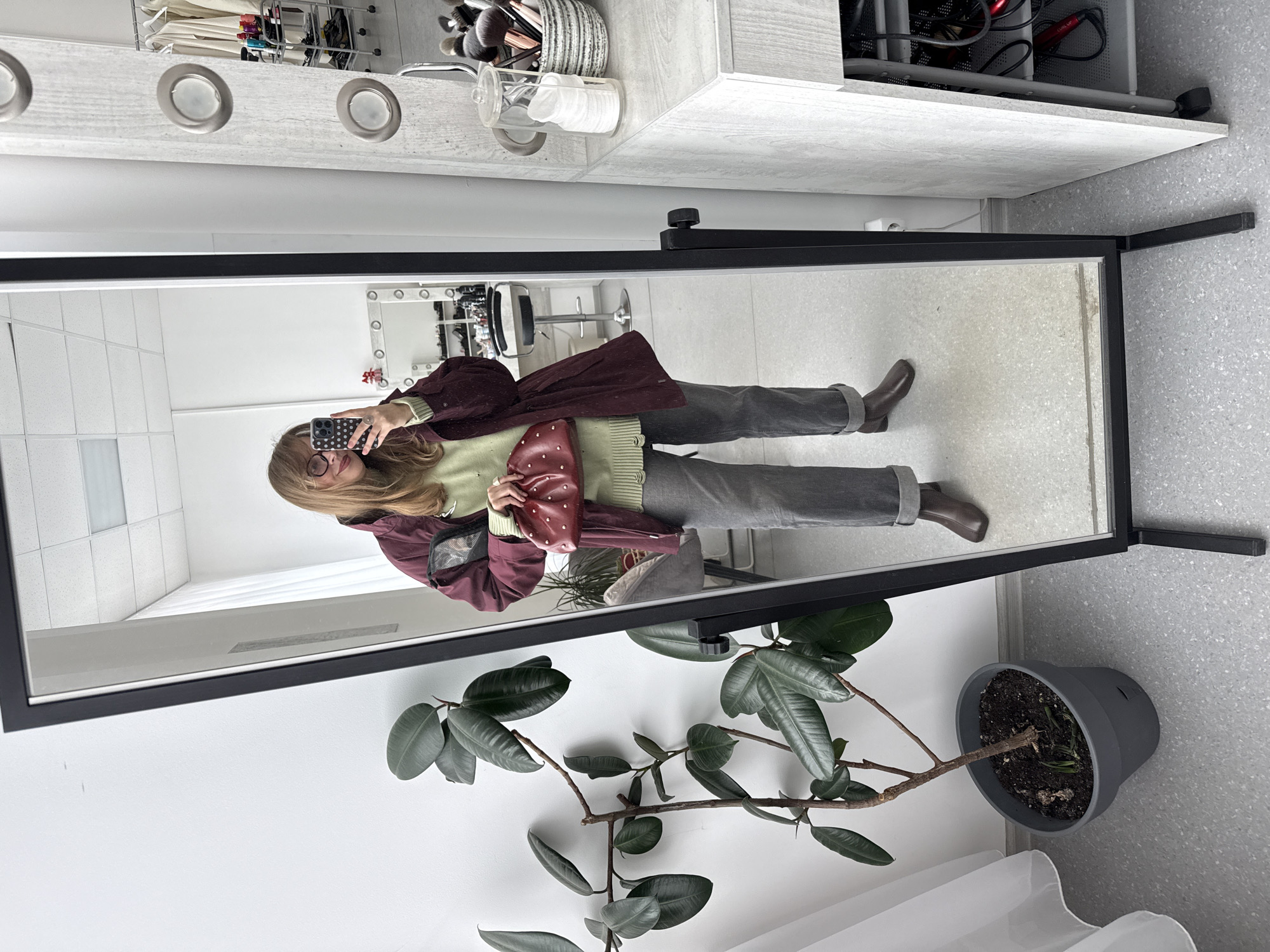 A woman is taking a photo of herself in a full-length mirror. The mirror is framed in black and stands on its own legs. In the reflection, she is wearing glasses, a light green sweater, gray pants, brown boots, and a burgundy jacket. She is holding a small, red, polka-dotted purse. Behind her in the reflection is a white wall with makeup lights, a makeup table, and a chair. To the right of the mirror is a potted plant with large, dark green leaves. The plant's pot is gray. The background wall is white, and the floor is a speckled gray. A section of white curtains is visible at the bottom of the image. The photograph appears to have been taken indoors. There is no indication of the location being in Izhevsk, Russia.