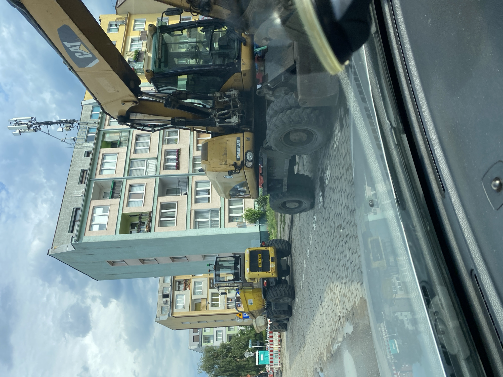The image shows a street scene in Szczecin, Poland, with two yellow excavators parked on a cobblestone street. The excavators are large construction vehicles, and one of them has the "CAT" logo visible. In the background, there are several apartment buildings, some of which appear to be under construction or renovation. The sky is cloudy, suggesting it might be an overcast day. The image appears to have been taken from inside a vehicle, possibly a car, looking out through the windshield, as indicated by the reflection on the glass. The excavators are the primary objects in the foreground and midground, and they are parked on a street that is wet in places, indicating recent rain or a damp surface. There are some barriers and what look like portable toilets further down the street, suggesting ongoing construction or an event. One of the excavators has a license plate with the text "BH67 DZR". The overall scene suggests a typical urban environment with construction activity.
