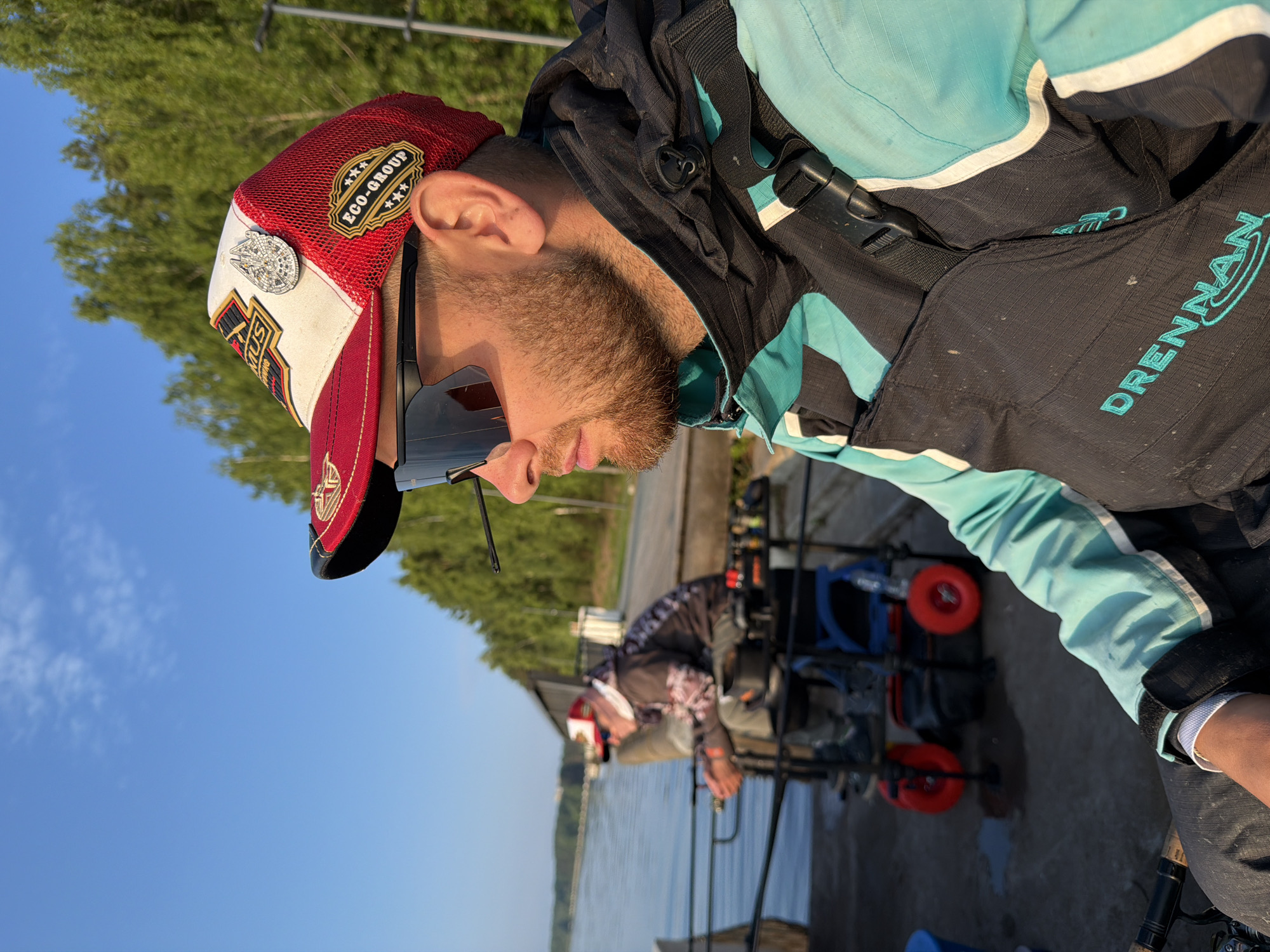 A man wearing a red and white trucker hat with patches, black sunglasses, and a turquoise and black fishing jacket is in the foreground, looking to the right. He has a beard and is sitting in what appears to be a fishing setup. Behind him, another person, wearing a red and white baseball cap, sunglasses, and a patterned hoodie, is seated in a fishing chair with red wheels. They are also facing away from the camera and towards the water. The background shows a body of water, a wooden pier or boardwalk, and a dense forest of green trees under a bright blue sky with some clouds. The scene is set outdoors, likely in a natural environment. The location is the city of Chaikovsky, Russia.
