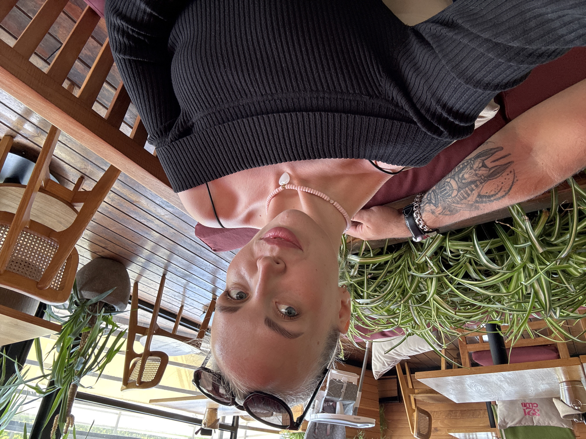 The image presents a tilted close-up view of a fair-skinned woman with grey eyes, sunglasses perched on her head, and wearing a black ribbed top and a pink beaded necklace with a white pendant. A man's arm with a large tattoo is visible near her right shoulder. The location appears to be an outdoor dining area with wooden furniture, potted plants, and views of what seems to be a city scene, likely in Sochi, Russia, based on the "321 CLUB" pillow visible in the scene.