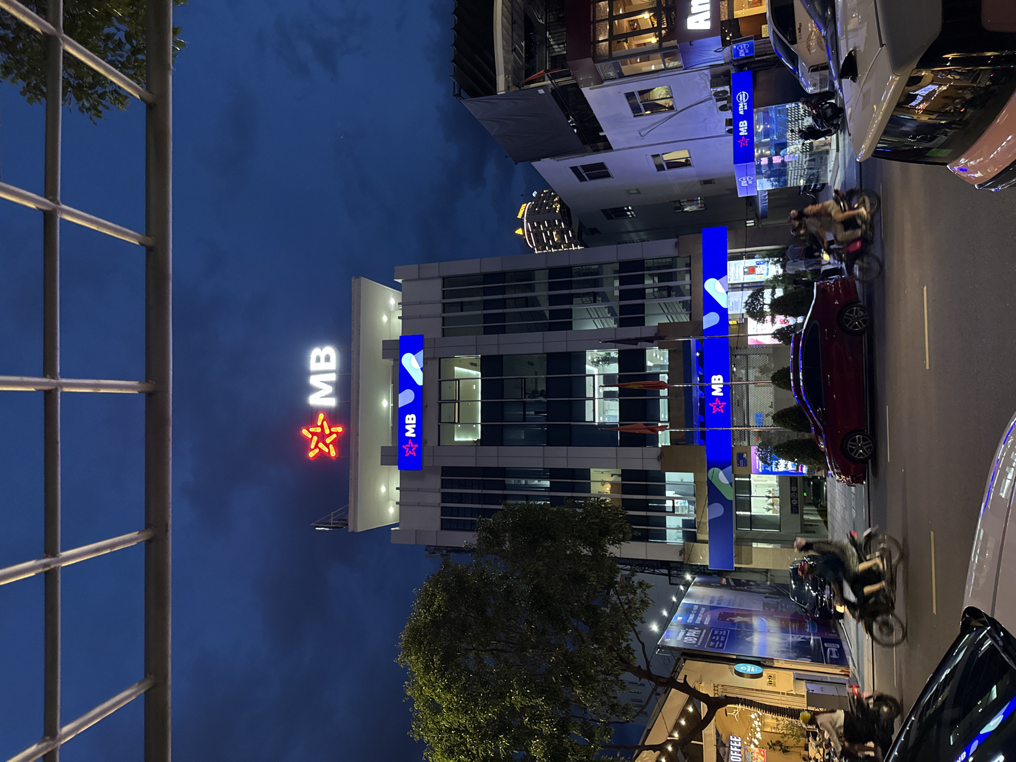 The image depicts a nighttime street scene in Nha Trang, Vietnam, likely in a commercial district. A multi-story building with prominent "MB" signage and a red star logo dominates the center, suggesting it's an MB Bank branch. The building features modern architecture with large glass windows and illuminated interiors. In the foreground, a street with traffic is visible, including several motorbikes with riders and at least two cars. One motorbike has two passengers, a driver and a pillion rider, both wearing helmets. A red car is stopped on the right side of the road, and a white car is partially visible in the bottom right corner. Other businesses with signage are visible on either side of the MB Bank building. A tree stands to the left of the MB Bank building, and a large advertisement billboard is also present. The sky is dark blue with some cloud cover, indicating dusk or night. The overall atmosphere is that of a busy urban environment at night.