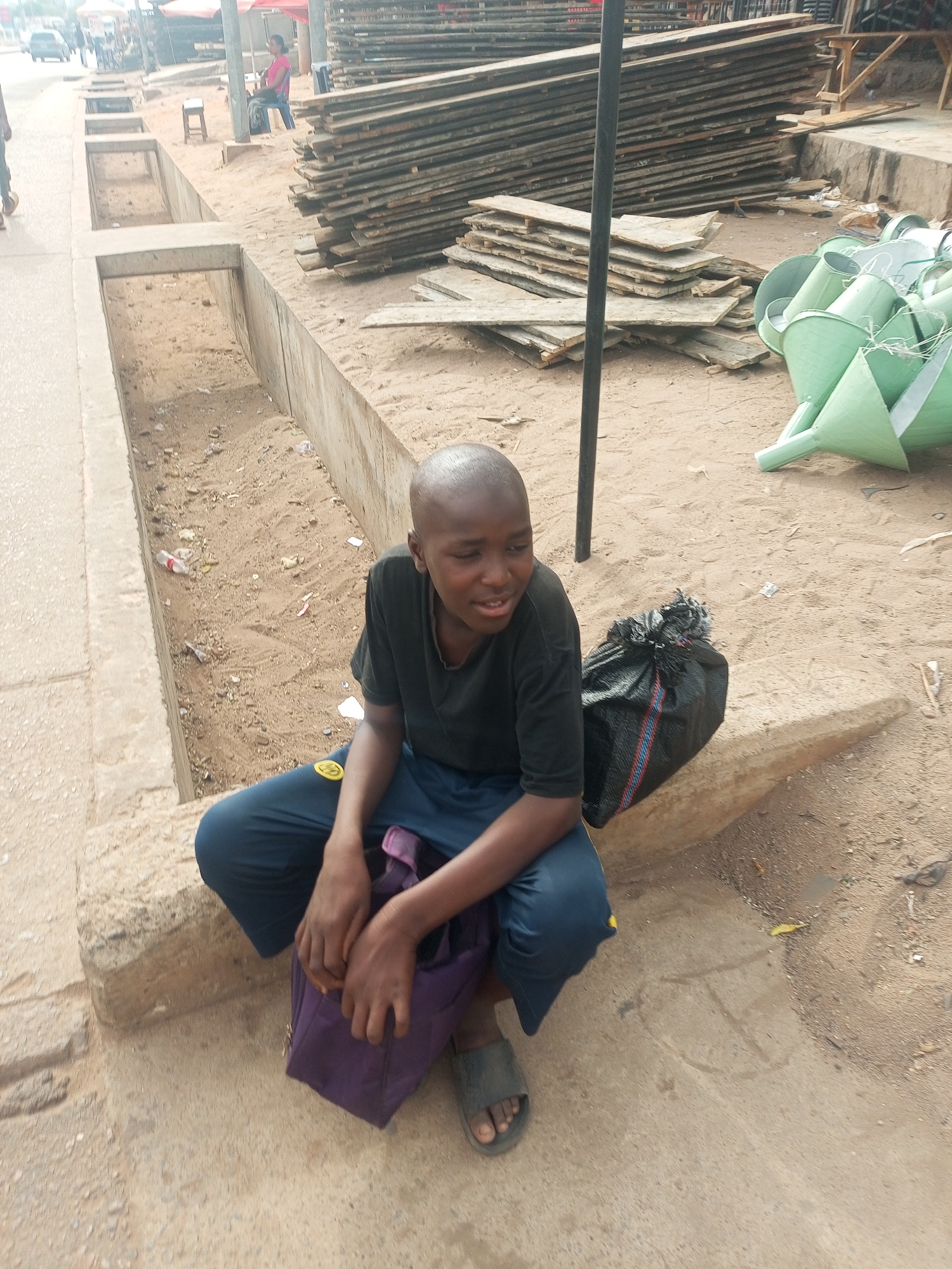 A young person sits on a concrete ledge alongside a street in Tempure, Nigeria. The individual wears a dark t-shirt, blue pants, and sandals. They have a shaved head and are holding a purple bag. Beside them is a black garbage bag.
In the background, there is a stack of weathered wooden planks, some green metal cones, and a concrete drainage ditch. Other people and structures are visible in the distance, suggesting an urban environment. The ground is mostly dirt and some pavement.