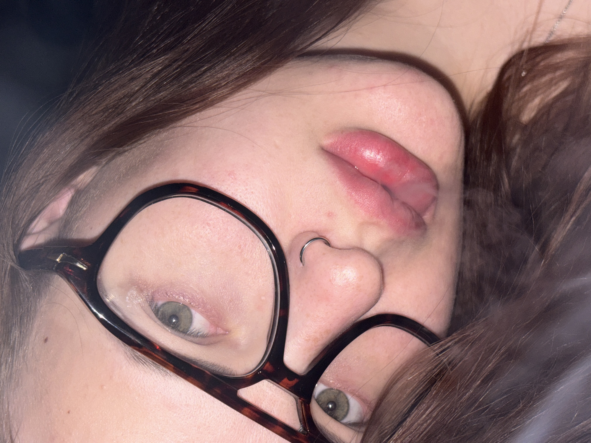 A close-up photograph depicts a person with light skin, brown hair, and glasses. They are positioned at an angle, with their face tilted towards the camera. The glasses have a dark brown tortoiseshell pattern. The person has a nose ring on the left side. Their lips are slightly parted. Their eyes are a hazel color. The background is dark and indistinct. There is no indication of Novomargaritovo, Russia.