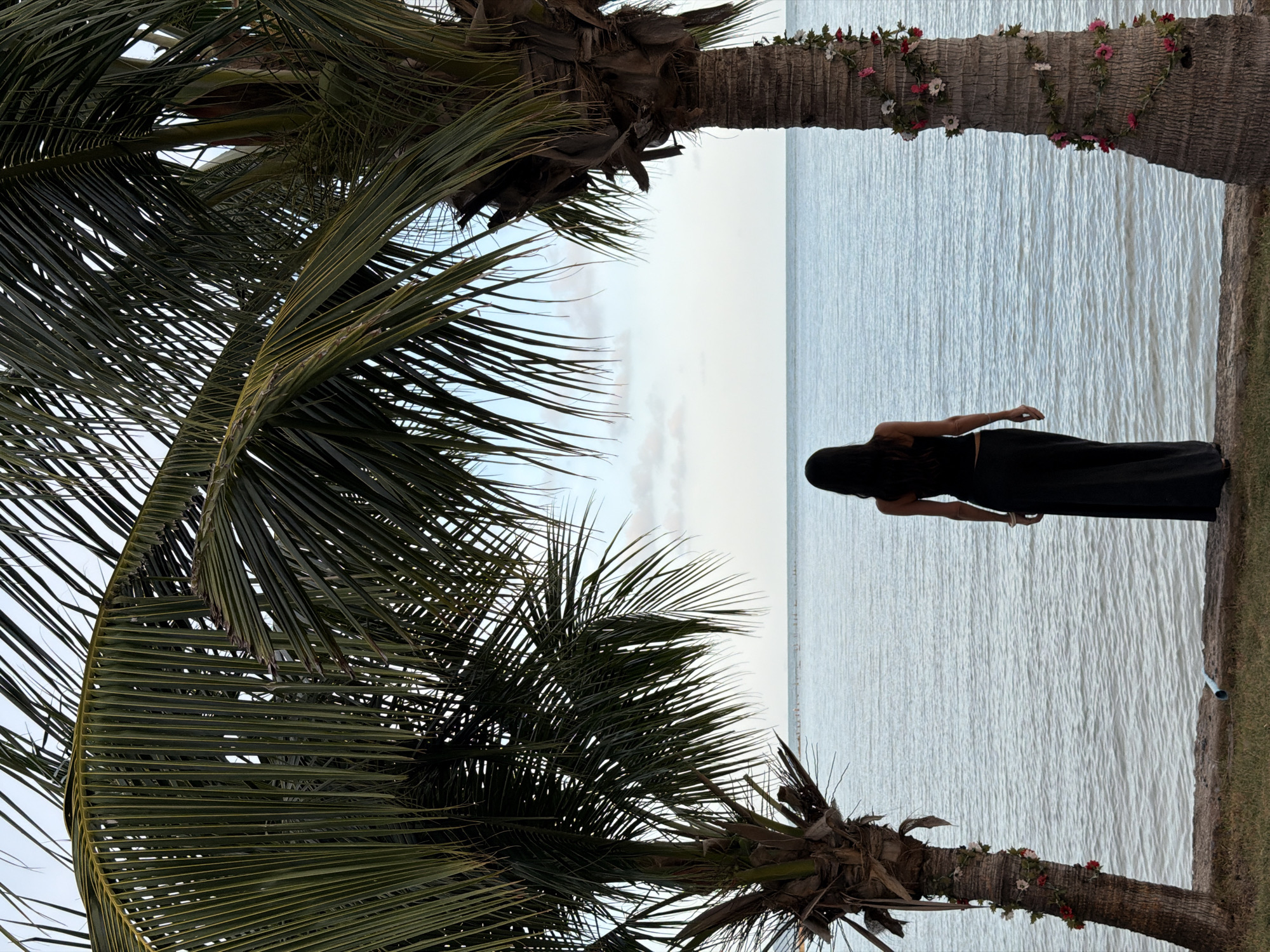 An overcast, daytime, medium shot captures a solitary woman standing on a grassy shore, her back to the camera. She is dressed in a black tank top and a long black skirt. Two palm trees, one on the left and one on the right, frame the scene, their fronds extending towards the sky and obscuring the upper portion of the image. The palm tree on the right is adorned with a garland of small pink and white flowers. The woman gazes out at a calm, wide expanse of light blue water. The horizon is visible in the distance, with a faint line of what appear to be structures or natural formations. The sky is a pale, hazy blue with scattered clouds. The overall impression is one of quiet contemplation against a tropical backdrop, possibly in the city of Ko Samui District, Thailand.