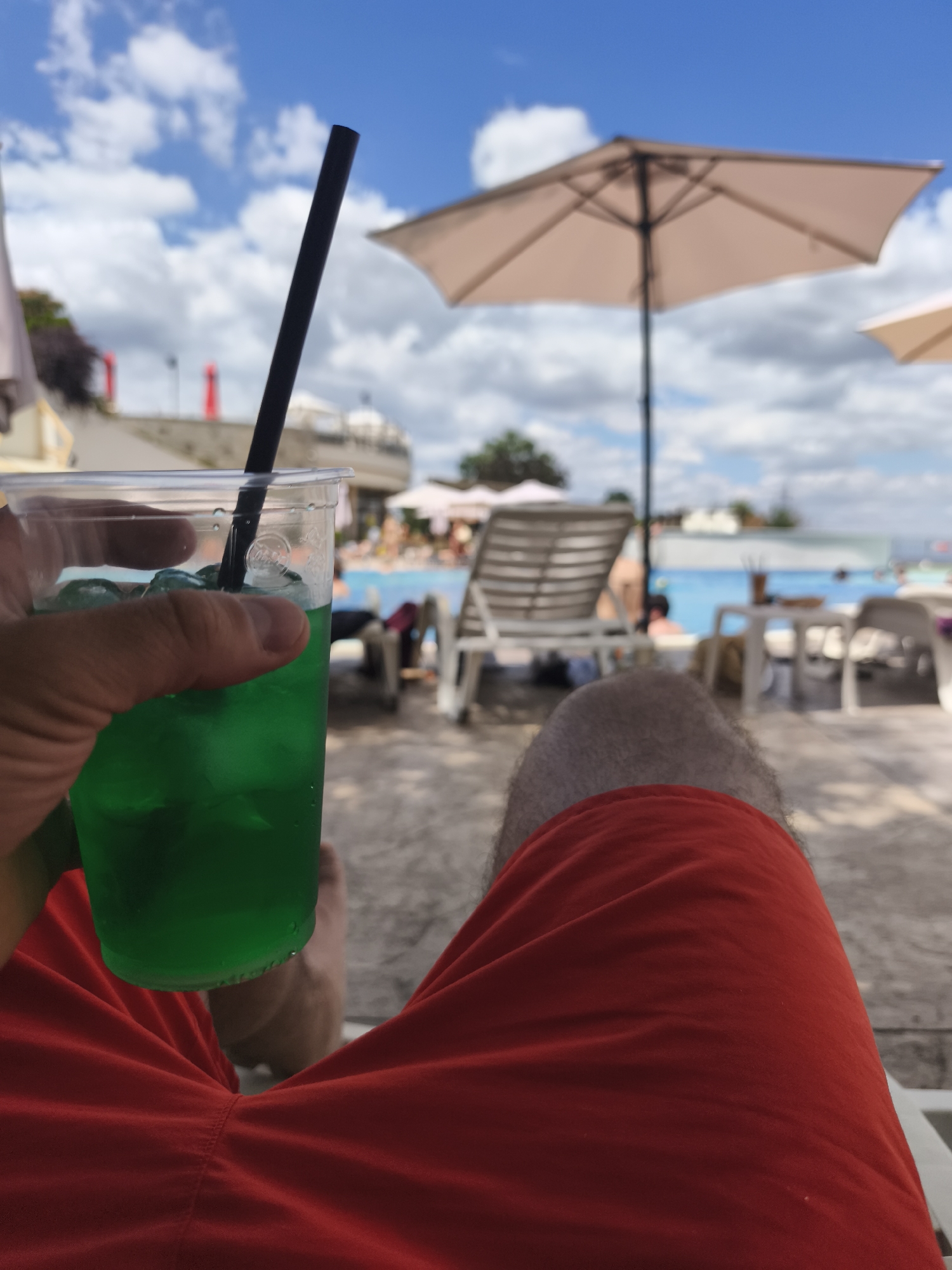The image shows a person's hand holding a plastic cup filled with a green liquid and ice, with a black straw. The person is wearing red shorts. In the background, there is a swimming pool area with lounge chairs, umbrellas, and people swimming and relaxing. The sky is blue with some clouds. The scene appears to be a resort or a hotel in Asenovgrad, Bulgaria.
