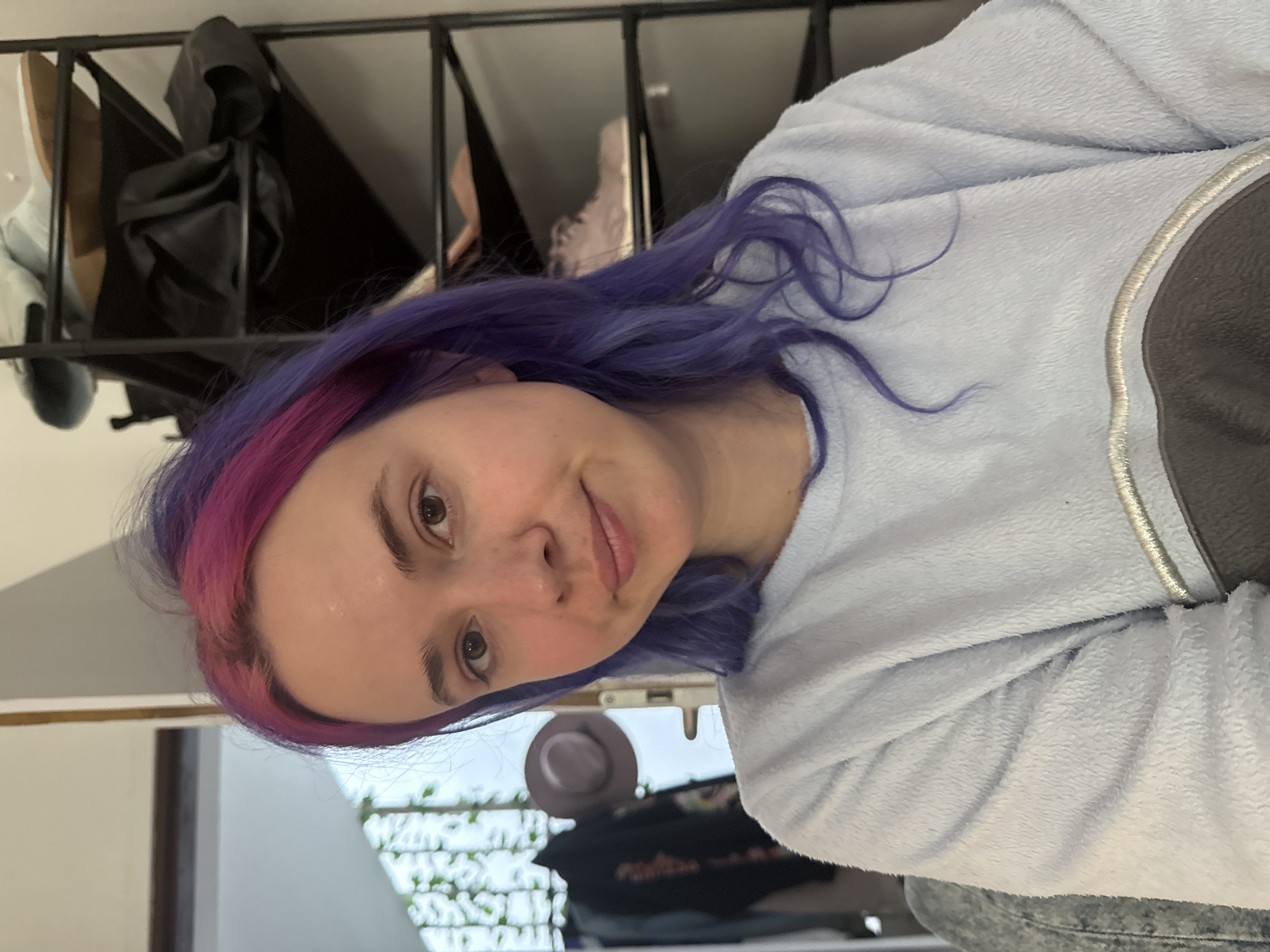 This indoor portrait shows a young person in the City of Zagreb, Croatia, looking directly at the camera with a gentle, subtle smile. The image is rotated 90 degrees counter-clockwise. The subject has distinctive long hair, which is primarily purple and features a vibrant pink section near the forehead. She has light skin and brown eyes.
She is dressed in a light blue, fuzzy long-sleeved top, possibly a fleece sweater, which has a circular grey patch with light stitching on the right chest area. A folded sleeve of the same material is visible, and she appears to be wearing grey bottoms, possibly jeans.
The background behind the subject is divided. In the upper left portion, a dark metal railing with vertical bars is visible. Behind the railing, various objects are stored, including a pair of light-colored shoes (possibly trainers) and a dark, possibly leather, bag or piece of clothing. The lower right background reveals a brighter space, likely an indoor area with natural light filtering in, possibly from a window or an open doorway. A white, patterned structure, possibly a railing or decorative screen, is visible through this opening, along with some greenery. A dark fedora-style hat rests on a surface in this area, and another blurred figure or dark garment can be vaguely seen in the deeper background. The lighting suggests it is daytime. There is no visible text in the image.