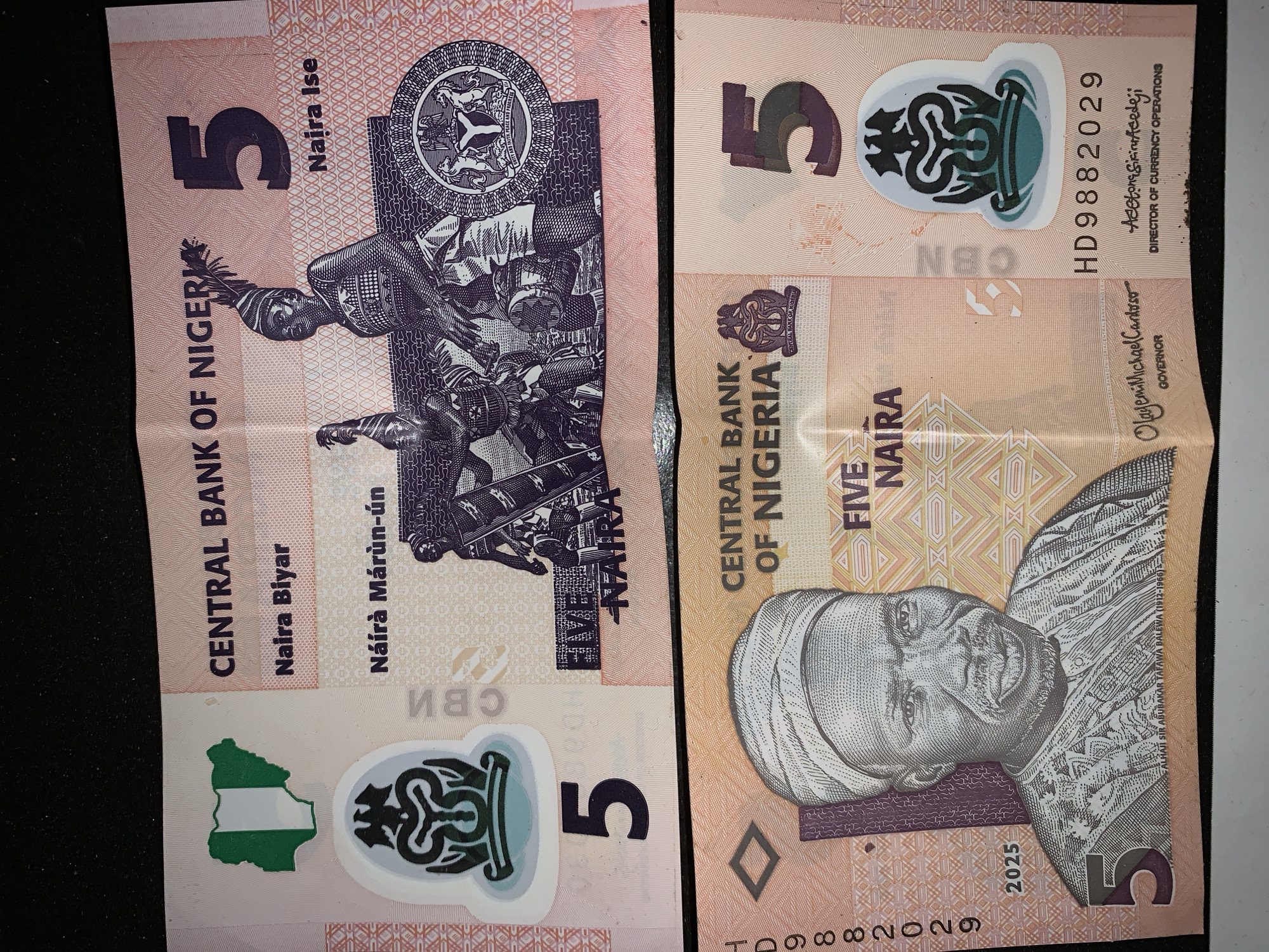 The image shows two Nigerian five naira banknotes. The left banknote features an illustration of people in traditional attire, a map of Nigeria, and the Central Bank of Nigeria's logo. The right banknote depicts Alhaji Sir Abubakar Tafawa Balewa, a former Nigerian Prime Minister. Both banknotes include security features like watermarks and serial numbers. The background is a dark, plain surface.