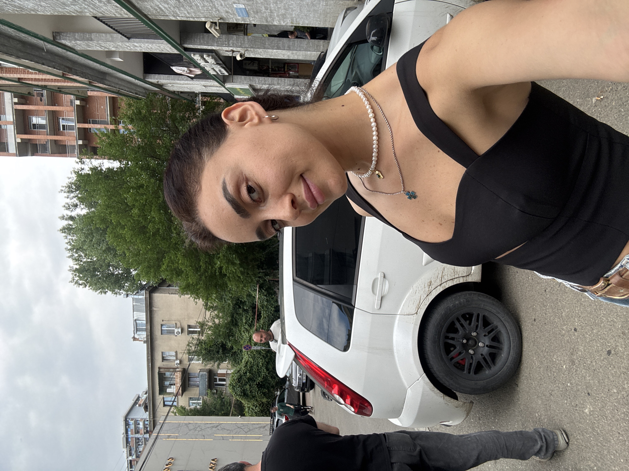 This media file shows a group of people outdoors in what appears to be a city street. The main subject is a woman in the foreground, taking a selfie. She is wearing a black top, a pearl necklace with a small blue pendant, and denim shorts. Her expression is relaxed and she is smiling slightly.
Behind her, a white car is parked on the side of the road. Other people are visible in the background, some standing near the car and others walking. The setting includes apartment buildings with balconies and trees. The sky is overcast, suggesting it might be a cloudy day. The overall scene gives the impression of a casual outing or gathering in an urban environment.
Visible text includes "PLAZA" on a building in the background. The location is identified as Tbilisi, Georgia.