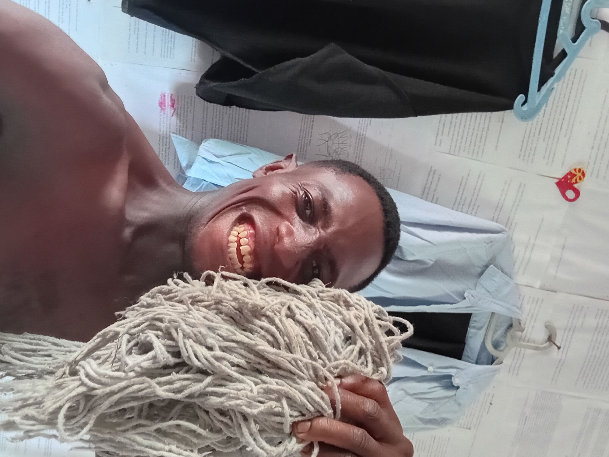 A shirtless Black male lies on his side holding a mop. He is smiling and looking at the camera. He is on a white surface with what appears to be printed text, a light blue shirt, and a black garment. A blue hanger and a small decoration are also on the surface. The location is potentially Akwete, Nigeria.