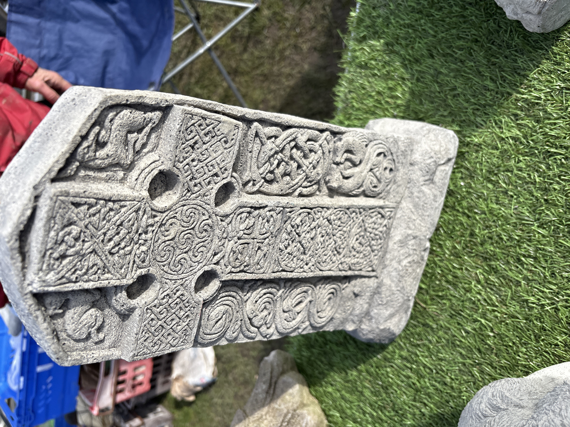 The image shows a grey, intricately carved Celtic cross lying on artificial grass. The cross is the main subject and features detailed knotwork patterns and possibly animal motifs. A portion of a person's red-sleeved arm and hand are visible in the upper left, along with a blue plastic crate and part of a metal folding chair. A partially visible stone is near the right side. The background is blurred, but suggests an outdoor setting with a mix of natural and artificial elements. The image was taken in Chirk, United Kingdom.