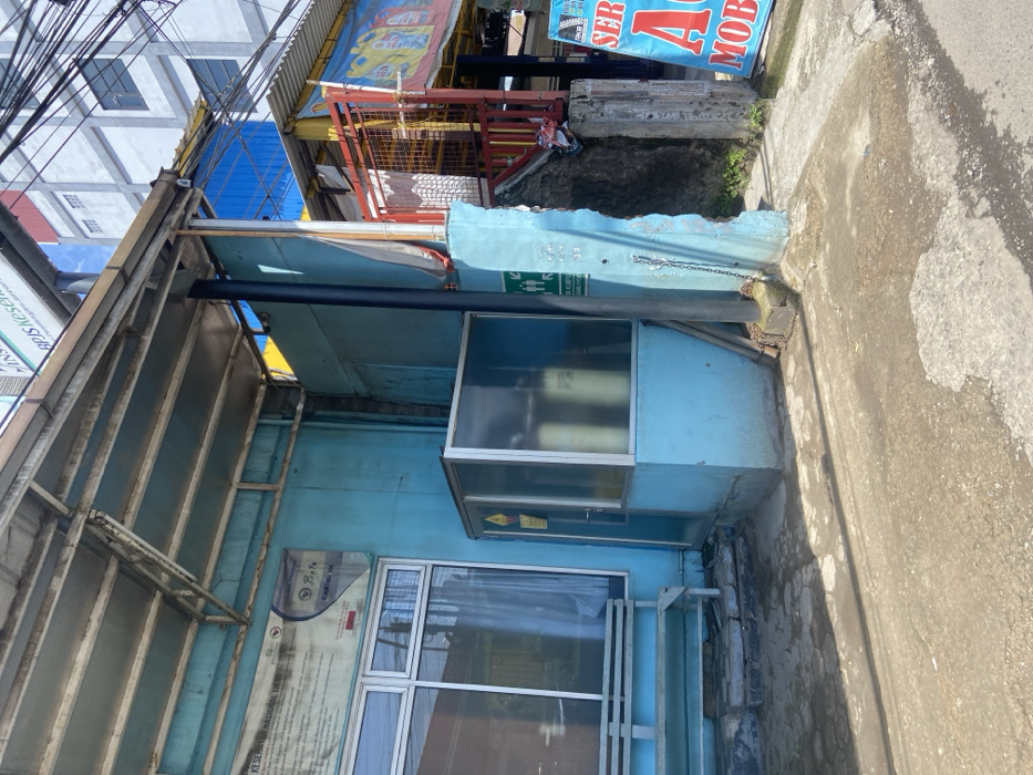 The image shows a small, partially enclosed area in front of a blue building, located in Padalarang, Indonesia, at coordinates -6.856 latitude and 107.498 longitude. The area is paved with cracked concrete, and there is a wooden bench on the left side. A blue, metal, glass-enclosed room with a door is directly in front of the bench. It appears to contain some sort of machinery. The room is set against a blue wall that continues around the corner. A metal chain hangs from the corner of the wall. The scene is brightly lit. There are no people in the image. There are wires and a partially visible building behind the blue building. There is a red gate on the right side of the image, and a sign that says "SERA MOB" is visible on the right side of the image.