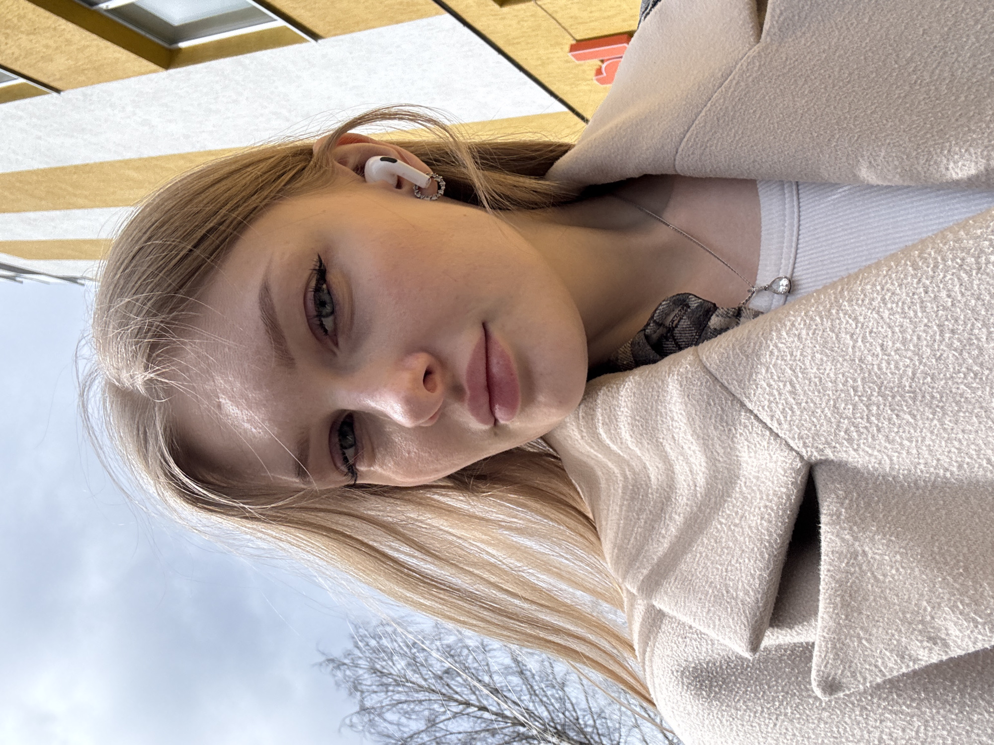 A fair-skinned young woman with blonde hair, wearing a white top and a beige coat, is pictured in an outdoor selfie. She has an AirPod in her left ear and a silver necklace. A building with yellow and white stripes is partially visible in the upper portion of the image. Bare tree branches can be seen in the lower left corner, suggesting winter or early spring. The location is inferred to be in Perm, Russia.