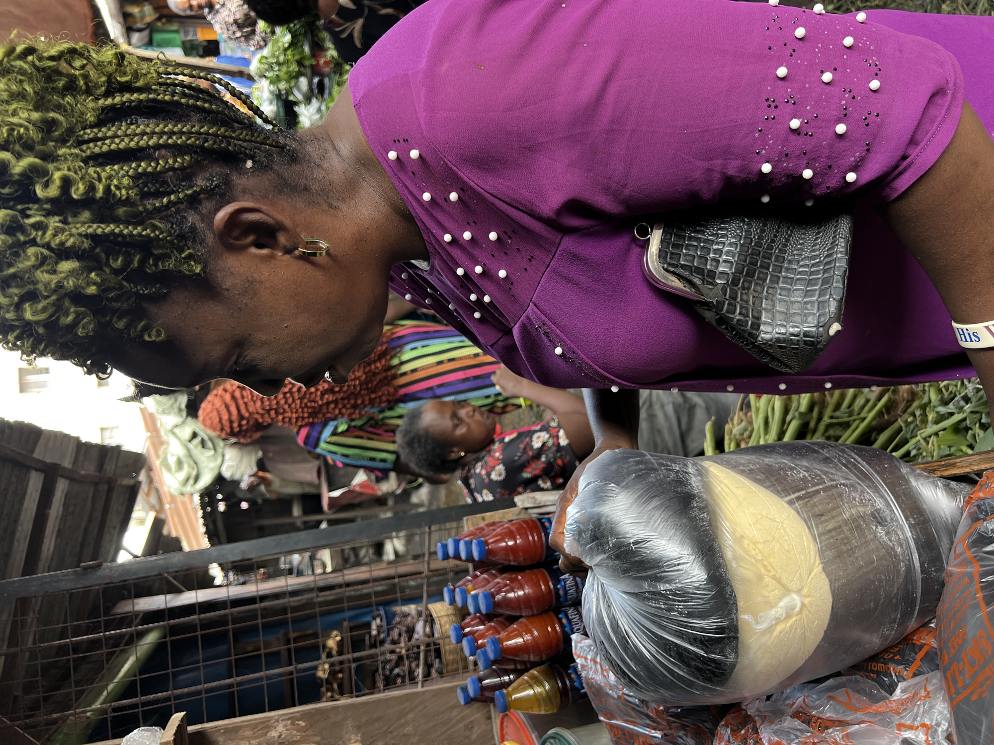 A woman with green braided hair wearing a purple dress with white and black embellishments is seen from the side, looking down. A black, crocodile-textured purse hangs from her shoulder. Several bottles of red liquid are visible, along with clear plastic bags containing yellow and black substances. In the background, another woman wearing a floral dress is lying down. The scene is within a market setting in Nkpor, Nigeria, indicated by the cluttered environment, the presence of various goods, and the metal fencing.