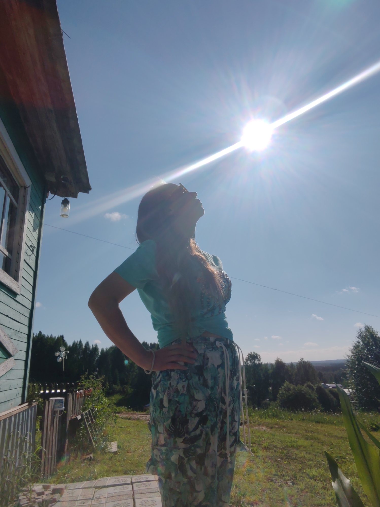 A low-angle, full-shot photograph captures a woman standing in front of a light blue wooden house with a worn, dark roof. She is positioned on a porch, indicated by the tiled pathway beneath her. The woman is silhouetted against a bright, sunlit sky, her face turned upwards and away from the camera. She has long, dark hair and is wearing a light blue t-shirt and a long, flowing skirt with a floral or leaf pattern in shades of blue and green. One hand rests on her hip, and the other is not visible. To her left, a portion of the house is visible, including a window with white trim. A glass light fixture hangs from the eaves of the house. In the background, a wooden fence, some vegetation, and a dense line of trees create a rural or suburban landscape. The sun is very bright, creating a strong lens flare that streaks across the upper portion of the image. The overall impression is a sunny, outdoor scene in a rural setting. The specific location is Чухлэм, Russia.