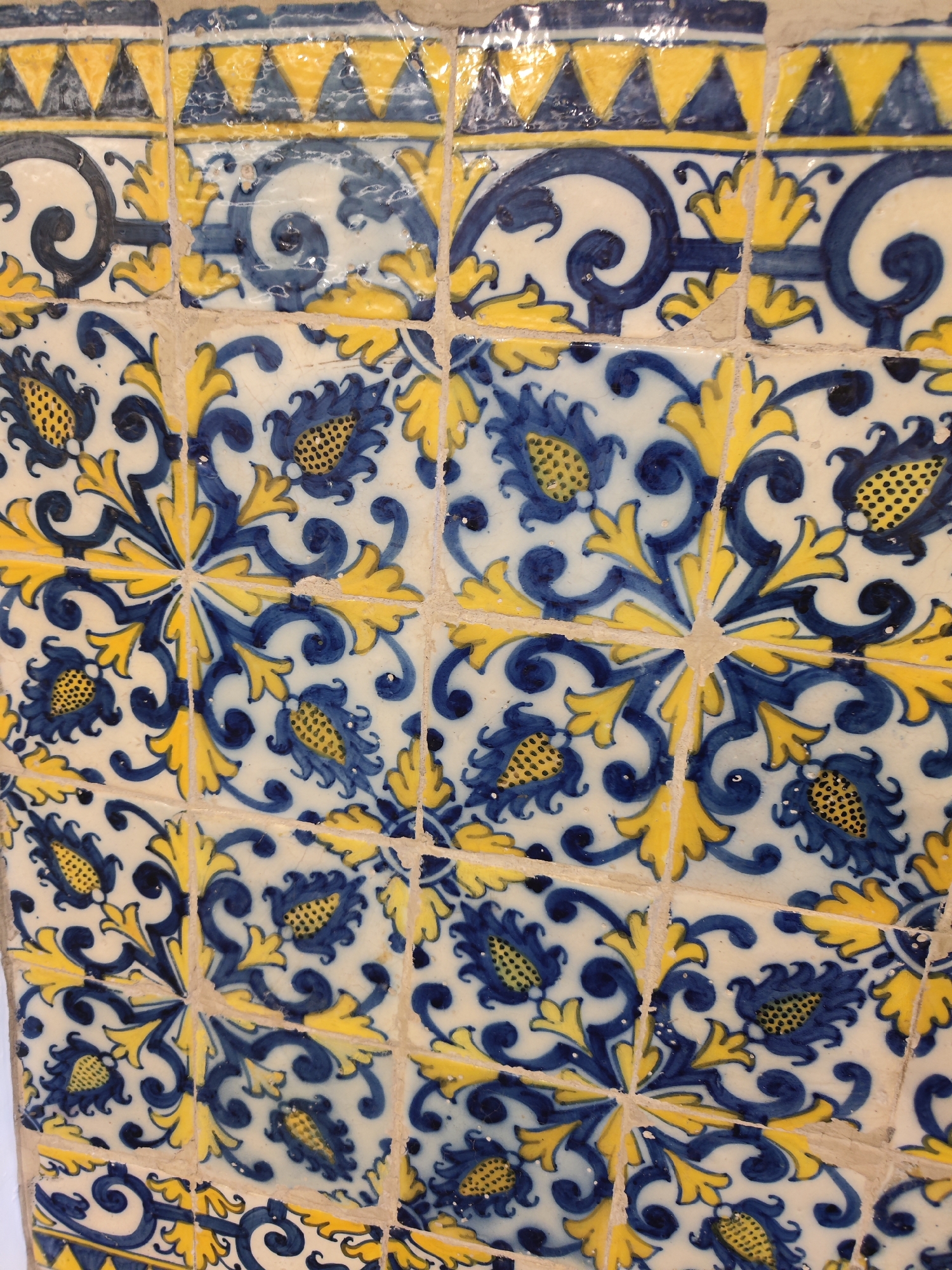 Close-up of a section of decorative tiles, likely azulejos, arranged in a repeating pattern. The tiles are predominantly white with blue and yellow floral and geometric designs. The upper part of the image shows a border of blue and yellow triangles. Below this, tiles depict stylized flowers in blue with yellow accents and a larger floral pattern in the center where four tiles meet. The grout lines between the tiles are thin and light in color. The tiles appear to be part of a larger installation, and the location is identified as Ponta Delgada, Portugal.