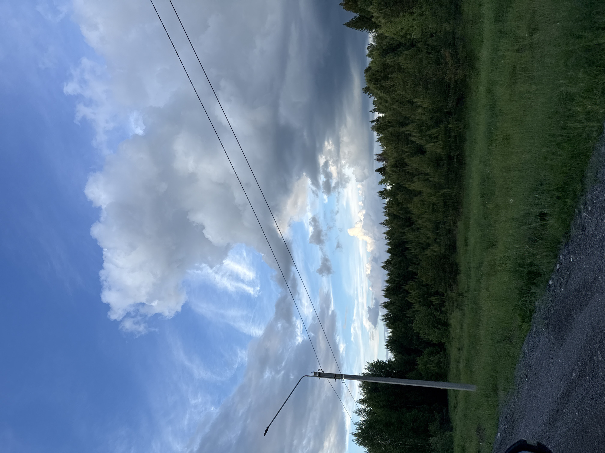 A landscape image shows an outdoor scene featuring a sky with varied cloud cover, a line of trees bordering a grassy area, and a section of road. In the foreground, the sky transitions from a deep blue with wispy clouds to a section with heavier, grey storm clouds. Utility lines run diagonally across the sky. On the right side of the frame, a dense line of trees separates the sky from a grassy field. The bottom right corner of the image shows a section of road surface, possibly gravel or asphalt. A streetlamp stands near the trees. The location is Марковский, Russia.