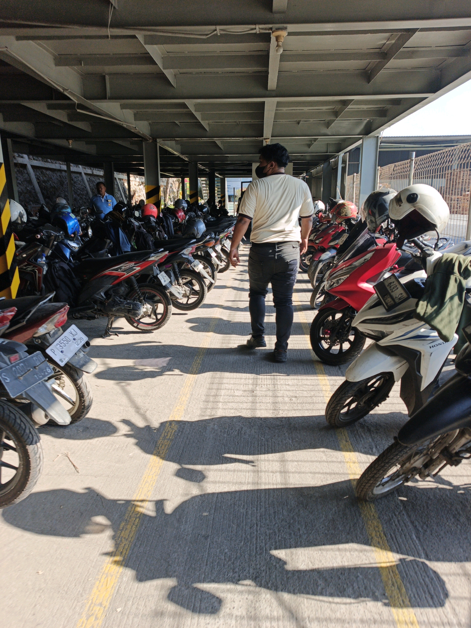 A man wearing a light-colored t-shirt and dark pants stands in a covered parking area for motorcycles. He is facing away from the camera, looking towards a line of parked motorcycles. The parking area has a concrete floor with yellow painted lines. The roof structure is made of grey metal beams and panels. Sunlight casts long shadows of the motorcycles and the man onto the ground. In the background, more motorcycles are visible, along with a person wearing a blue uniform. The scene suggests a typical day in a motorcycle parking lot, possibly at a commercial or public facility in Ngoro, Indonesia.