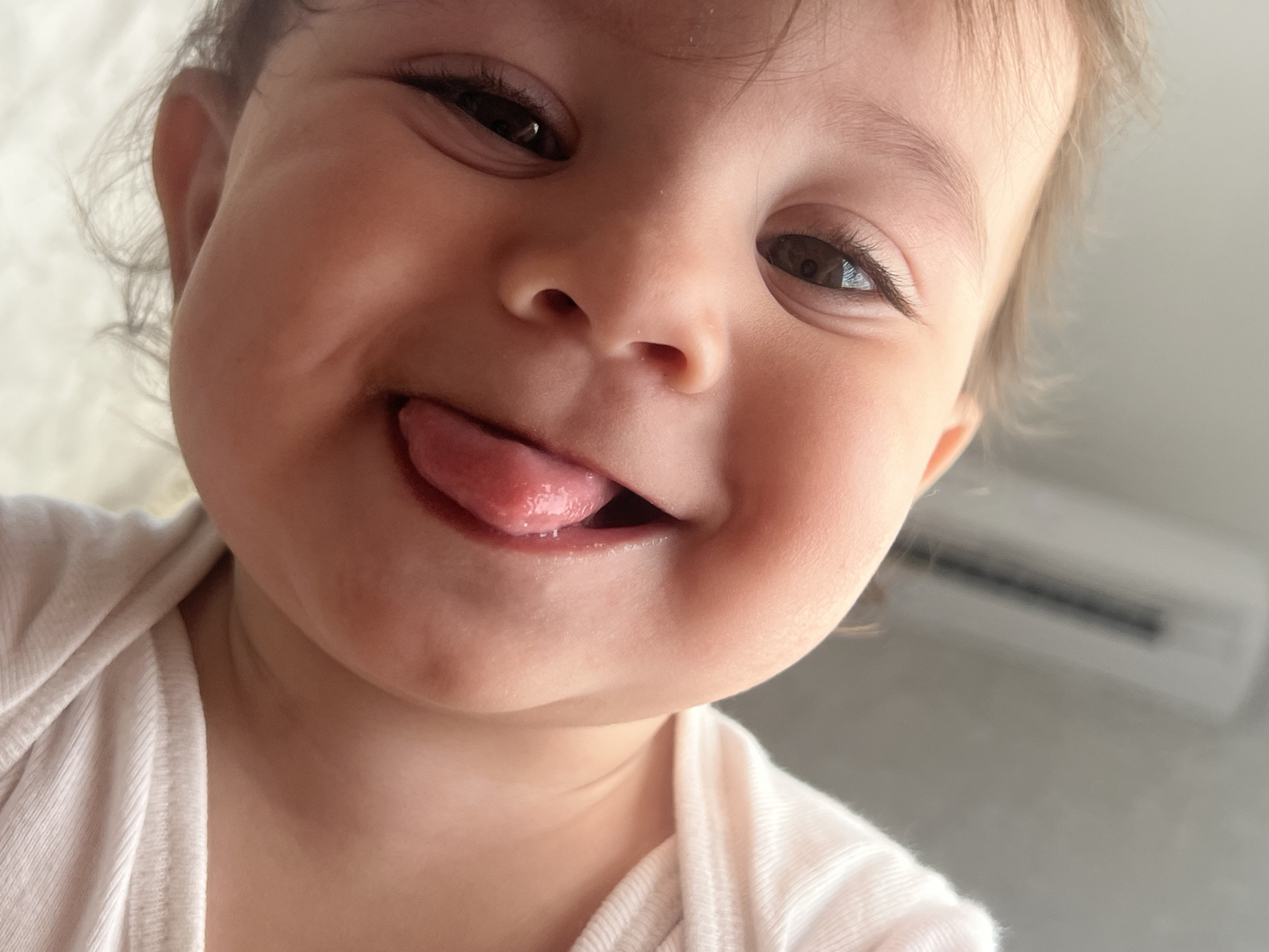 A close-up, low-angle photograph captures a joyful baby, likely a toddler, indoors in Elena, Bulgaria. The child's face dominates the frame, looking down towards the camera with a playful and happy expression. The baby has wide, dark eyes and light, wispy hair visible around its face and ears. Its mouth is open in a wide grin, with its tongue playfully sticking out, suggesting a moment of childish mischief or fun.
The baby's skin tone is warm, and its cheeks appear full and soft. It is wearing a light-colored, possibly white or cream-colored, ribbed shirt or onesie, visible around its shoulders and neck. The background is softly blurred and appears bright, indicating an indoor setting, likely during the daytime. On the upper right side of the image, a portion of a white air conditioning unit with dark horizontal vents is visible, out of focus. No text is visible in the image. The overall impression is one of innocence, happiness, and playful curiosity.