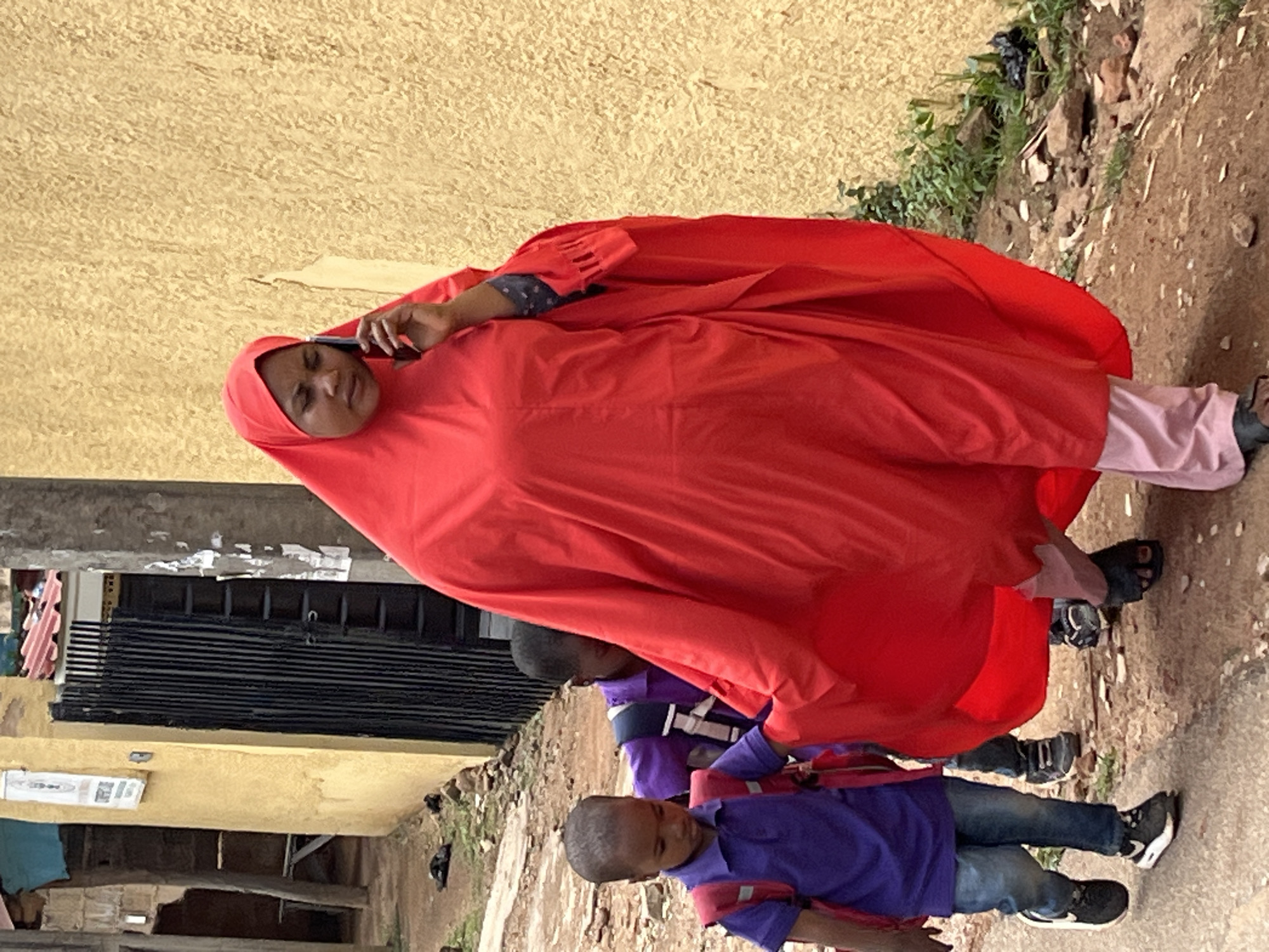 The image depicts a street scene in Jos, Nigeria, during the daytime. The central figure is a woman, facing towards the viewer, wearing a bright red full-length garment and a matching red hijab covering her head. She holds a black smartphone to her right ear, appearing to be engaged in a phone call. Her expression is neutral.
Behind and to the lower right of the woman, two young boys are visible. The boy in the foreground wears a purple long-sleeved shirt, dark jeans, and black sneakers with white soles, and carries a red backpack. His face is not fully visible. Another boy, partially obscured by the woman's red garment, also wears a purple shirt and is positioned further back. The children appear to be walking along a dirt path.
The ground is uneven, composed of dirt, small rocks, and some scattered debris. On the right side of the path, there is sparse green foliage and larger rocks, suggesting an uncultivated area adjacent to the path. In the background, a building with a light beige or yellow textured wall is prominent. Towards the left of the image, behind the subjects, a structure with horizontal black slats, possibly a gate or security barrier, is visible. On the lower section of the yellow wall, there's a white rectangular object with indistinct black text, likely a poster or sign. The overall scene suggests everyday activity in a semi-urban or residential setting.