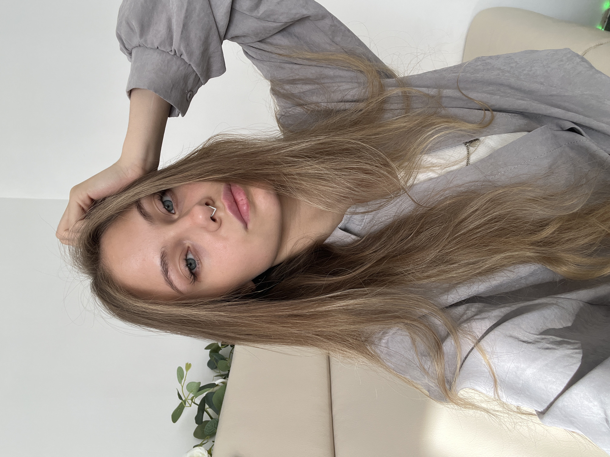 A young woman is posing for a picture. She is wearing a gray jacket and a small nose ring. Her long brown hair falls across her face and body. She is resting her hand on her head and looking directly at the camera. The background includes a white wall and a beige couch with some greenery. The location Фоки, Russia, cannot be confirmed from the image.