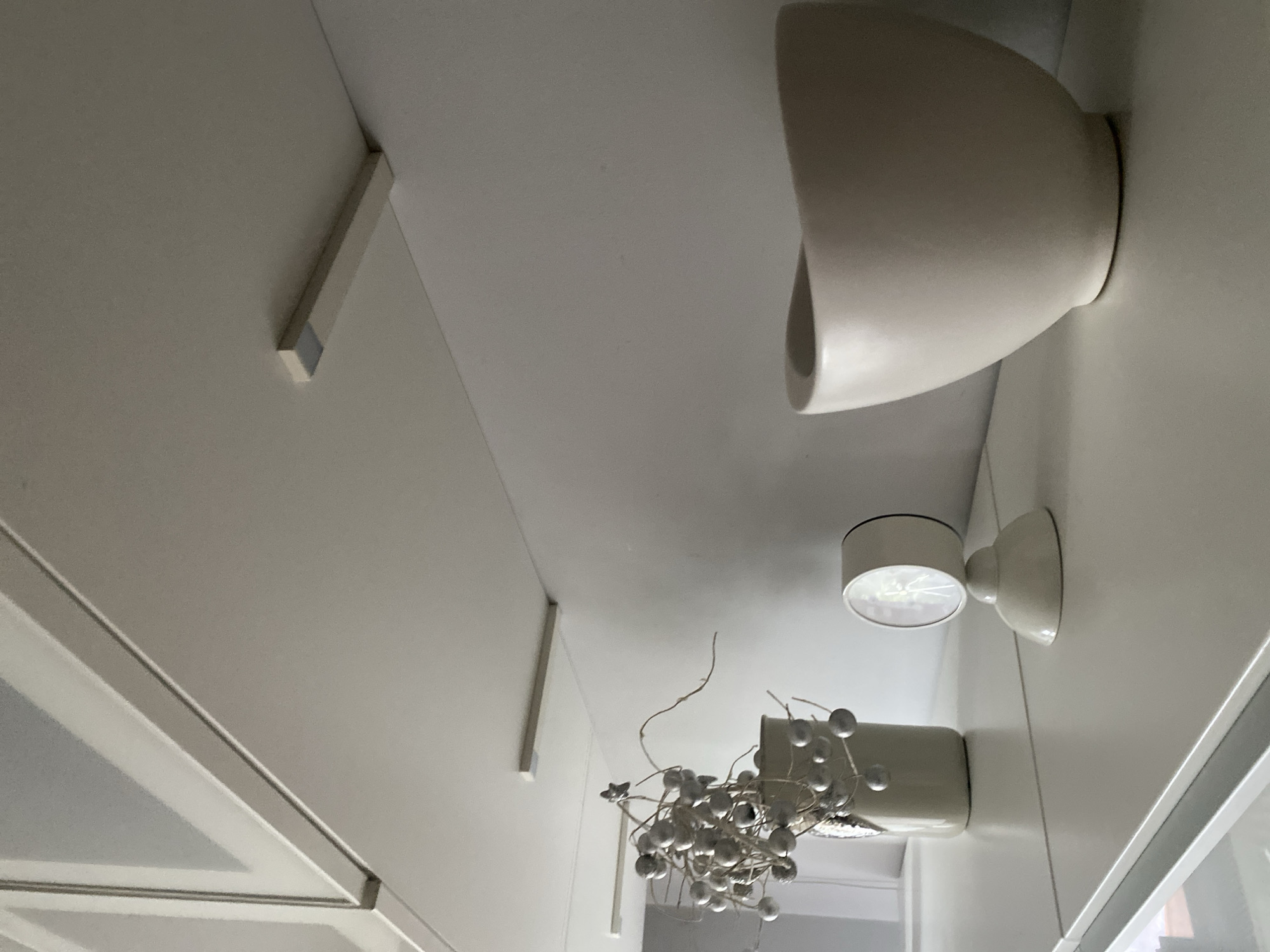 The image depicts an interior scene with white shelving units. On the shelves, there are decorative items including a vase filled with silver spherical ornaments, a small white clock, and a beige bowl. Two rectangular light fixtures are mounted on the underside of the upper shelving unit. The image does not contain information that would allow for the determination of its location in Wrocław, Poland, or any other specific city.