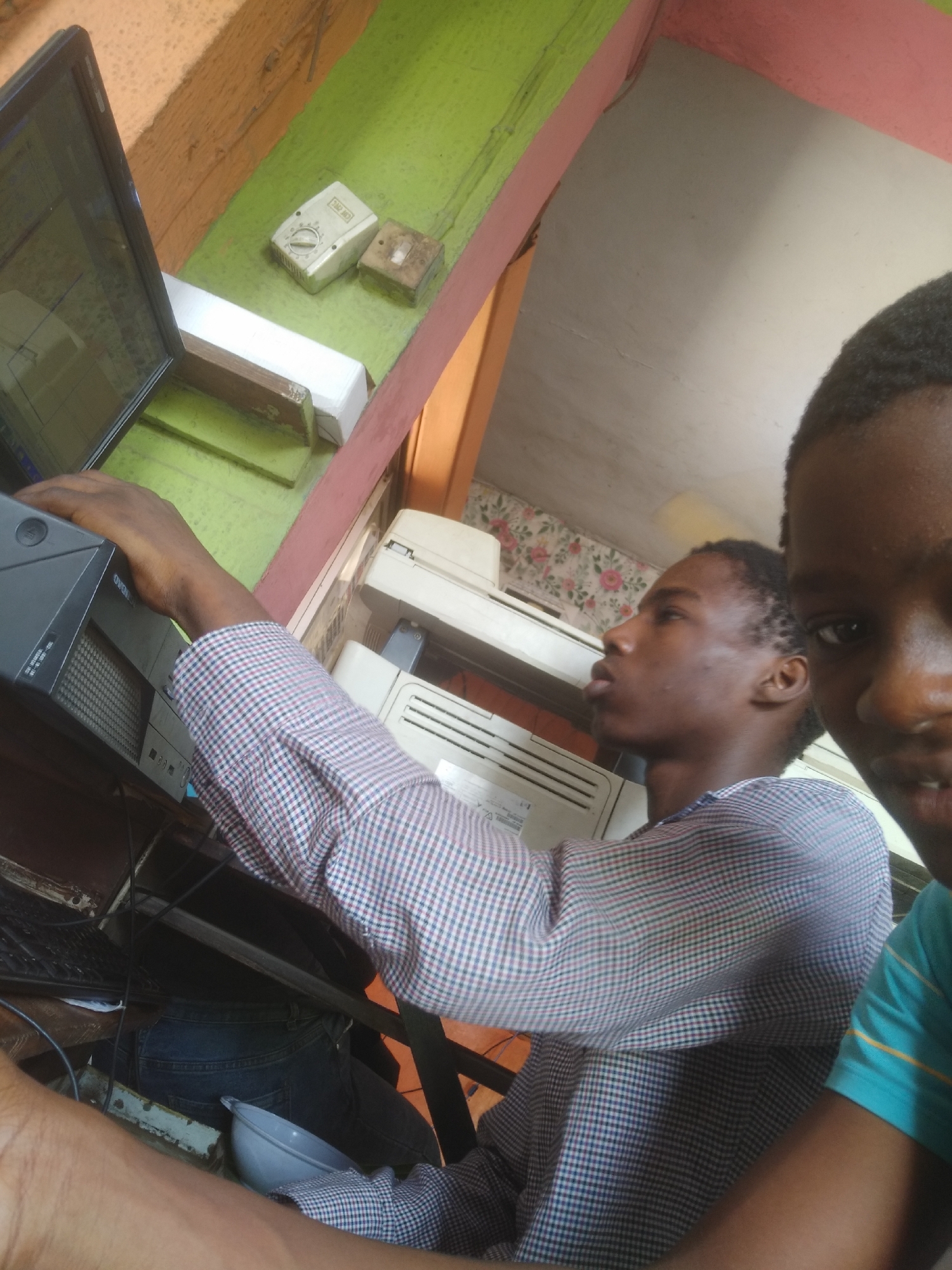 The image shows two young men in a room in Shangisha, Nigeria. One man is in the foreground, wearing a blue shirt. The other man is seated and working at a computer. He is wearing a checkered shirt. The room contains a desktop computer, a printer, and some electrical fixtures. The walls are painted in various colors.