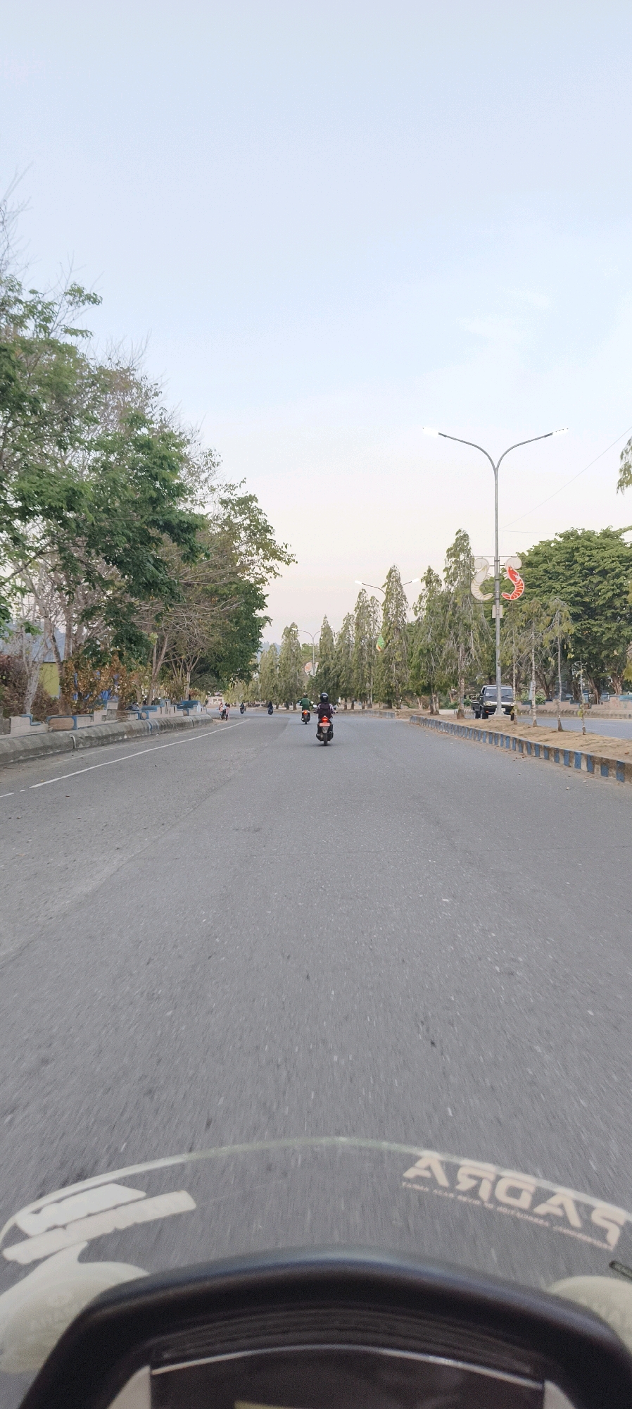 The image captures a view of a road in Waisai, Indonesia, looking forward from the perspective of someone riding a motorcycle. The road is paved and appears to be a multi-lane thoroughfare. On the left side of the road, there are trees and some low-lying vegetation, as well as a gray concrete barrier with blue trim. On the right side of the road, there is a median strip lined with tall, slender trees and decorative streetlights with ornamental attachments. Several motorcycles are visible on the road, some with riders. One car is parked on the right side of the road. The sky is clear and a light, pale blue, suggesting it is either early morning or late afternoon. The foreground of the image shows the handlebars and windshield of a motorcycle, with some text visible on the windshield.