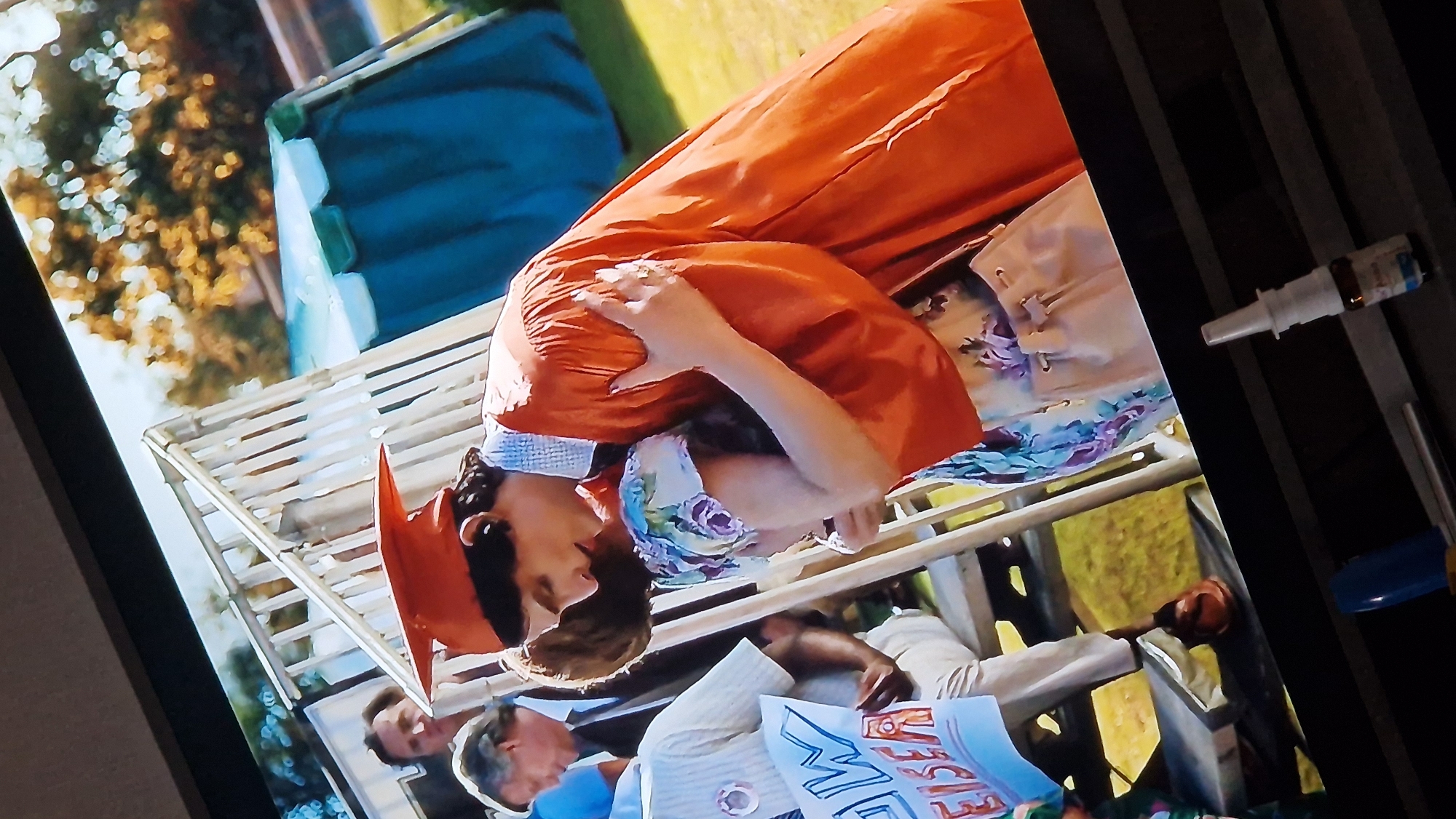 In the city of Unknown, Unknown, a graduation ceremony is taking place outdoors, possibly in a schoolyard or park. The scene is captured from a slightly elevated angle, showing a group of people in the foreground and middle ground, with trees and a blue tent-like structure in the background.
In the immediate foreground, a young person wearing an orange graduation cap and gown is embracing another person. The graduate's gown is vibrant orange, and they are wearing a light blue and white patterned dress underneath. The other person being embraced is mostly obscured, with only their head and shoulder visible. They appear to be wearing a dress with a floral pattern of purple and blue flowers. The graduate's arms are wrapped around the other person, and they are leaning in for a kiss or a hug.
To the left of the main interaction, a person is seated, wearing a light-colored collared shirt and light-colored pants. They are holding a sign that says "NOW EISER" in blue and red lettering. Another person, a man with dark hair and a suit jacket, is standing behind them, looking towards the graduate. Further in the background, more people are visible, and a white railing structure suggests a set of bleachers or stairs. The ground is covered in green grass. A large, blue structure with a white top, possibly a tent or canopy, is visible behind the crowd, and trees with yellow and orange foliage suggest it might be autumn.
The overall atmosphere of the image appears to be emotional and celebratory, with the embrace and the graduation setting indicating a significant milestone.
