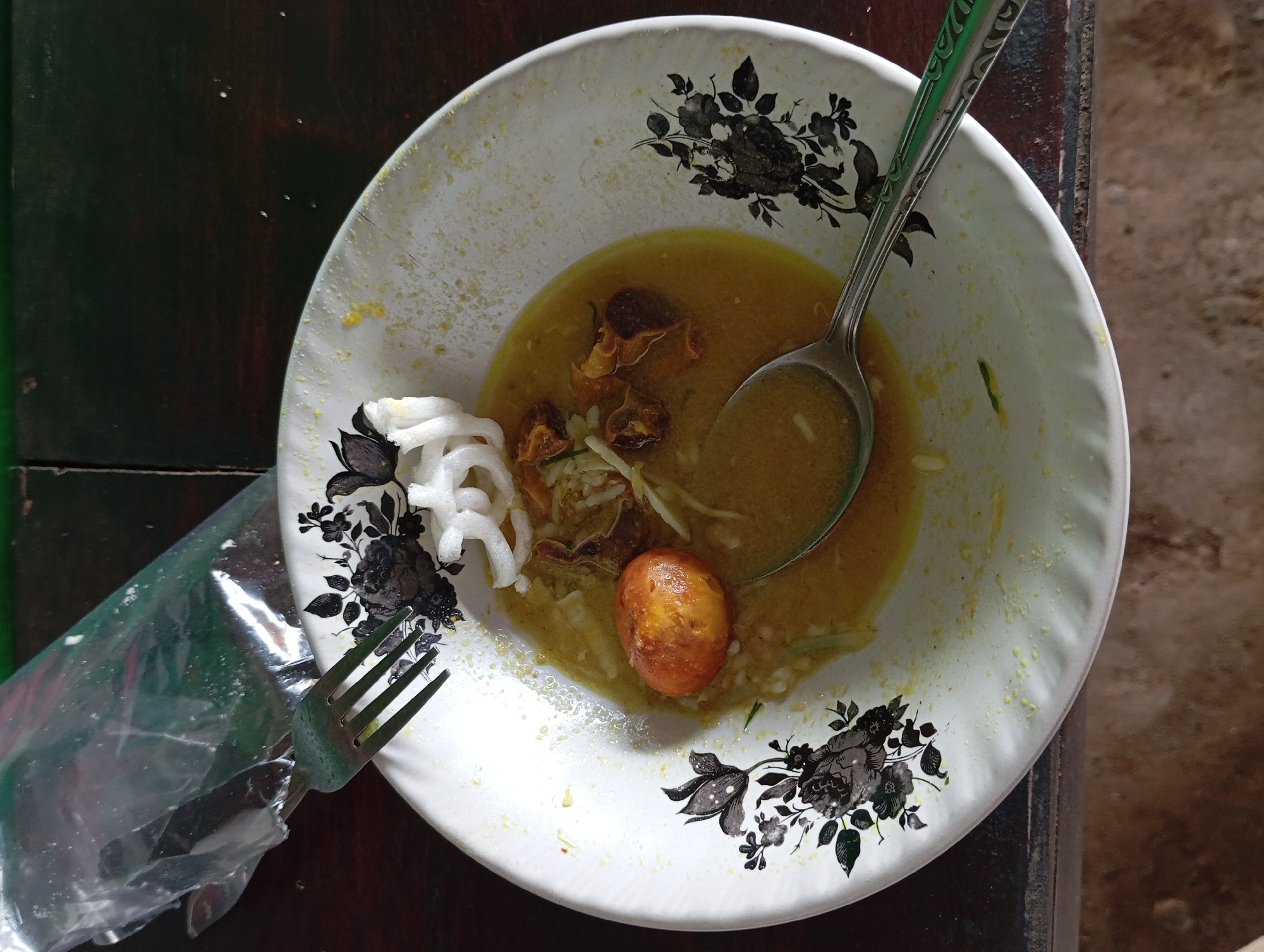 A top-down, medium shot captures a partially eaten meal on a white, floral-patterned plate. The plate contains a yellow broth with visible pieces of meat and a whole boiled egg. A spoon rests in the broth, and a fork is positioned over the rim of the plate, near a crumpled plastic bag. The meal is set on a dark, wooden surface. The scene appears to be in Lemahbang, Indonesia, suggesting a casual dining setting.