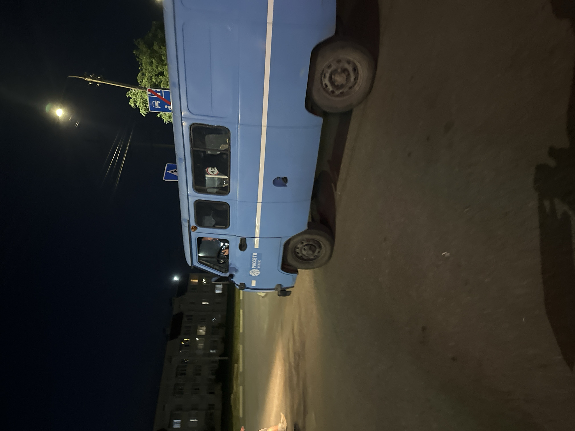 In the city of Nevinnomyssk, Russia, at night, a blue utility van with white stripes is parked on an asphalt road. The van has "РОССЕТИ" written on its side, indicating it belongs to a Russian energy company. A speed limit sign showing "40" is visible through the van's window. In the background, a multi-story apartment building with illuminated windows suggests a residential area. Streetlights illuminate the scene. The image captures a moment of stillness, with no apparent immediate action beyond the presence of the van and the distant building. The overall environment appears to be a typical urban setting at dusk or night.