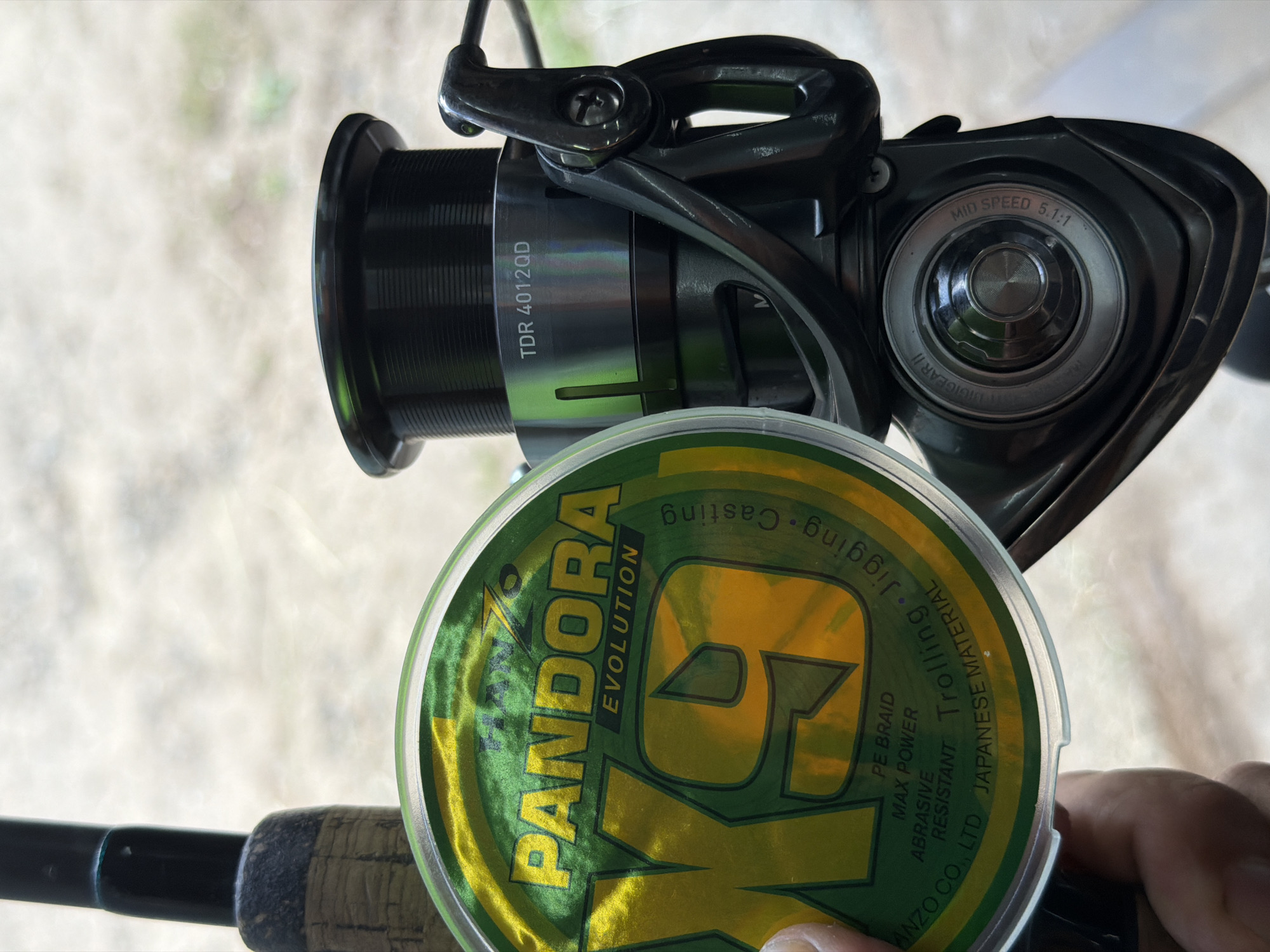 The image displays a close-up of fishing equipment, set outdoors in what appears to be Chaikovsky, Russia. The main subjects are a black and silver fishing reel, a spool of green and yellow fishing line, and a section of a fishing rod with a cork handle. A person's hand is visible holding the spool of line, suggesting an activity related to preparing for fishing, such as spooling line or inspecting gear.
The background is brightly lit and blurred, indicating a daytime setting with natural outdoor surroundings, possibly showing ground or foliage. The lighting is diffused, perhaps from an overcast sky or dappled sunlight.
Visible text on the fishing reel includes "TDR 4012QD", "MID SPEED 5.1:1", and "DIGIGEAR II" (partially visible). The spool of fishing line prominently features "PANDORA EVOLUTION" and a large "G" design. Additional text on the line spool reads "PE BRAID", "MAX POWER", "ABRASIVE RESISTANT", "Trolling • Jigging • Casting", and "ANZO CO., LTD JAPANESE MATERIAL".