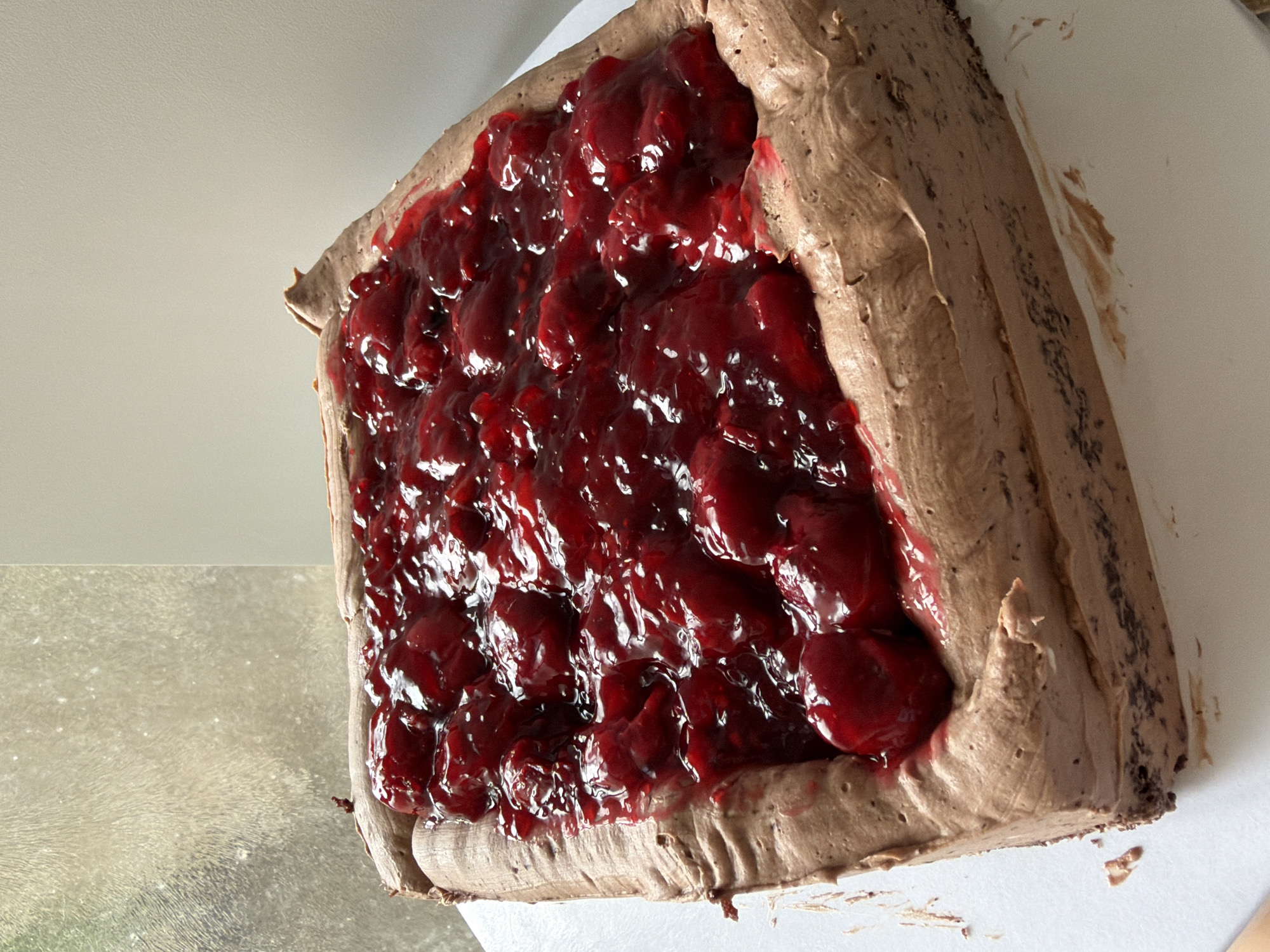 A close-up shot presents a square slice of chocolate cake topped with a thick layer of bright red cherry filling. The cake slice is placed on a white surface. The frosting is a milk chocolate color. The surface beneath the cake appears to be a reflective window or tabletop. The location is not evident from the visual context.