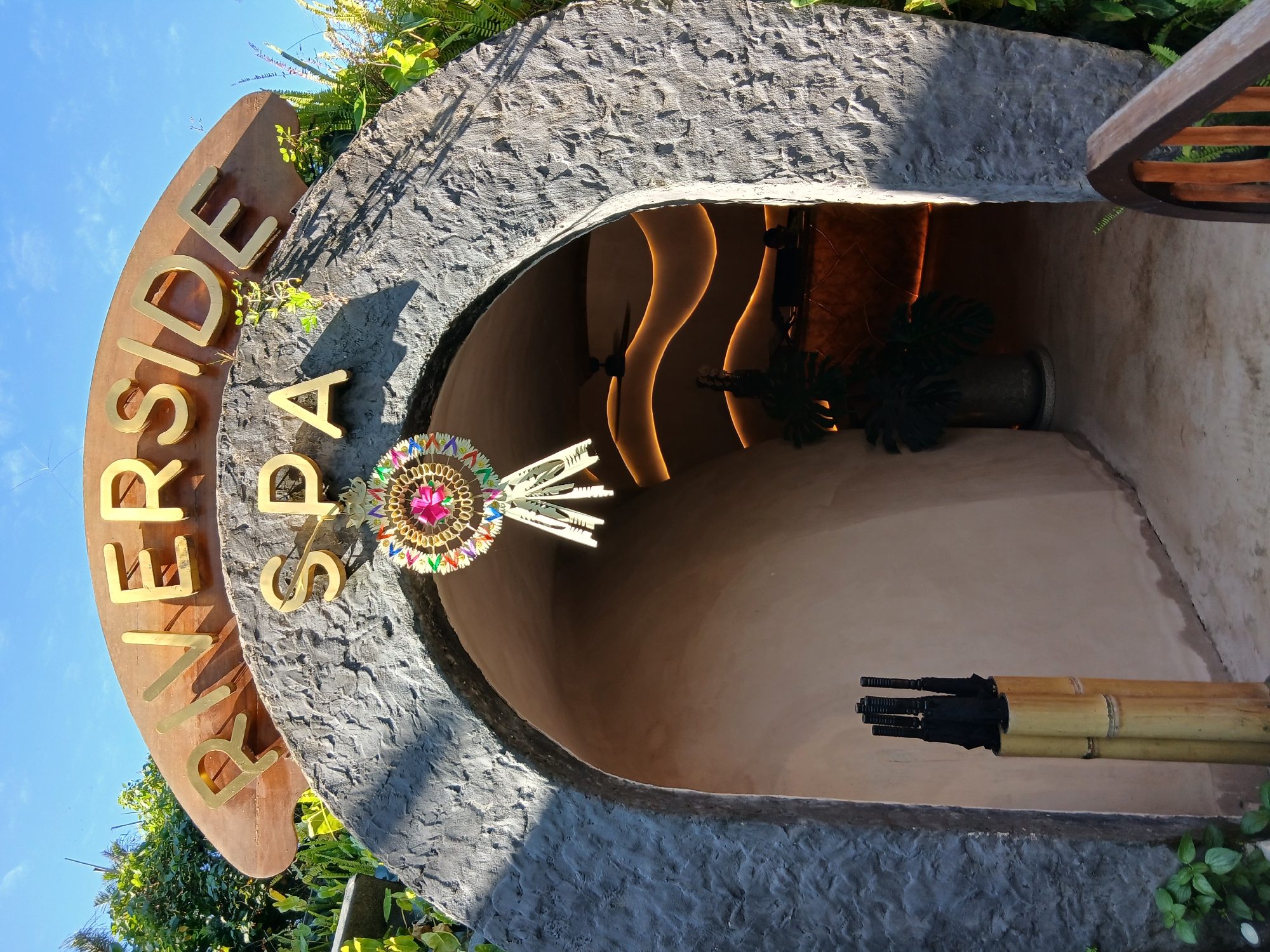 The image shows the entrance to "RIVERSIDE SPA" in Kediri, Indonesia. The entrance is an arched opening in a rough, stone-like structure, overgrown with greenery. Above the arch, golden letters spell out "RIVERSIDE SPA" on a curved wooden sign. A decorative hanging ornament, featuring a pink jewel and colorful woven elements, is suspended from the arch. To the left of the entrance, a bamboo stand holds several black umbrellas. Inside the spa, a dark hallway is visible with wavy, illuminated walls and a ceiling fan. A potted plant and what appears to be a reception counter are also present in the dimly lit interior. The sky is blue with some clouds.