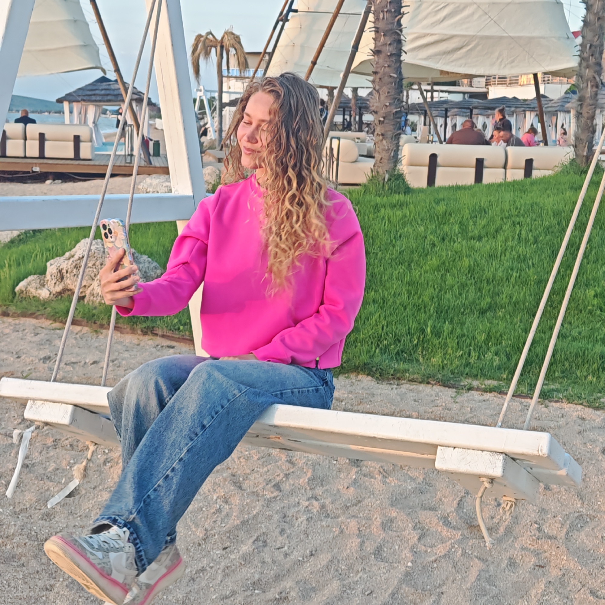 A woman sits on a white wooden swing taking a selfie with her phone. She is wearing a bright pink sweatshirt and blue jeans. Her shoes are predominantly clear with white accents and pink soles. The ground around the swing is sand. In the background, there is green grass, palm trees, and covered seating areas with white cushions. The scene is located in городское поселение Щелкино, Ukraine.